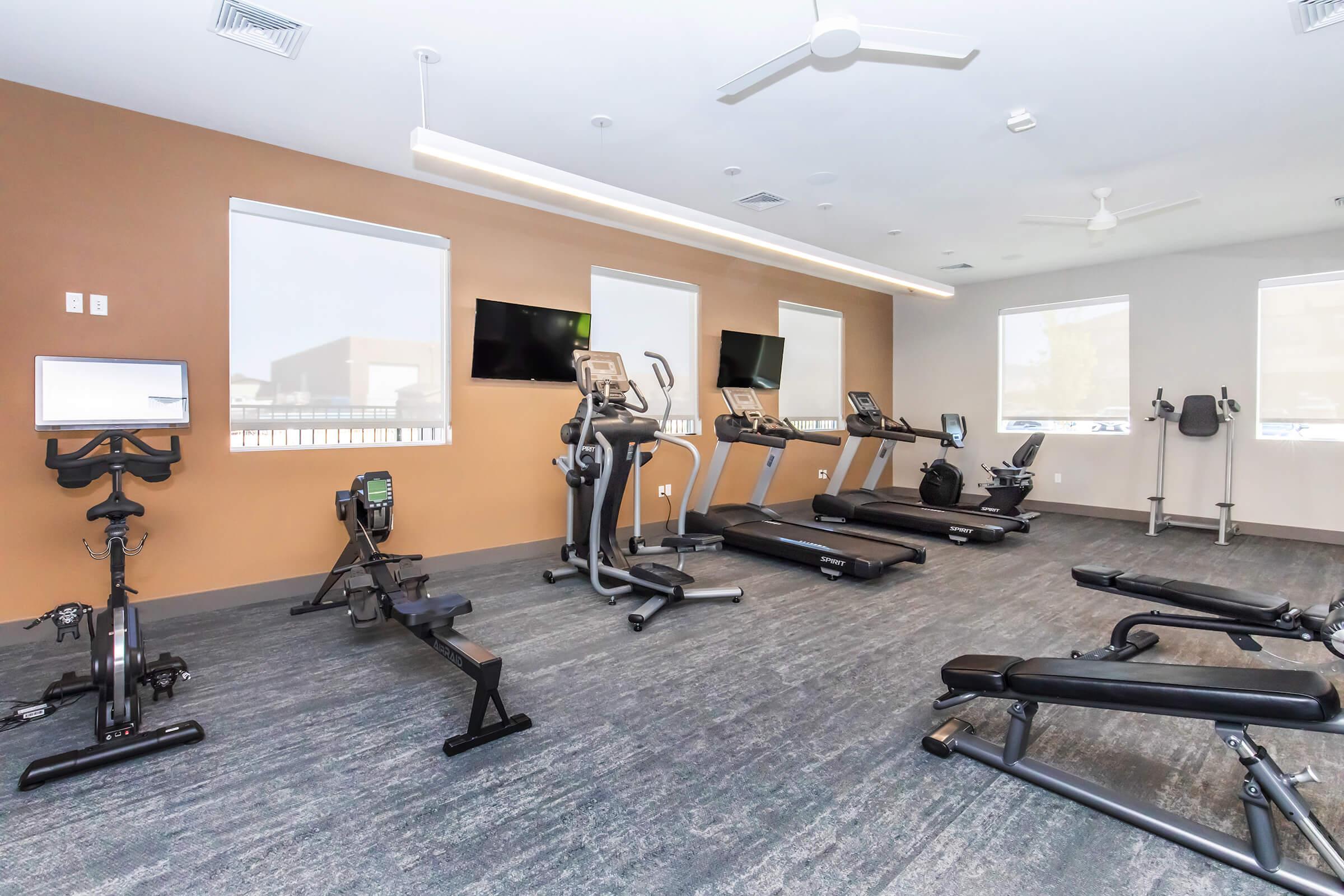 A modern gym interior featuring various exercise equipment, including treadmills, a rowing machine, stationary bike, free weights, and a multi-gym station. The room has large windows, ceiling fans, and a neutral color scheme with a brown accent wall, creating a bright and inviting workout space.