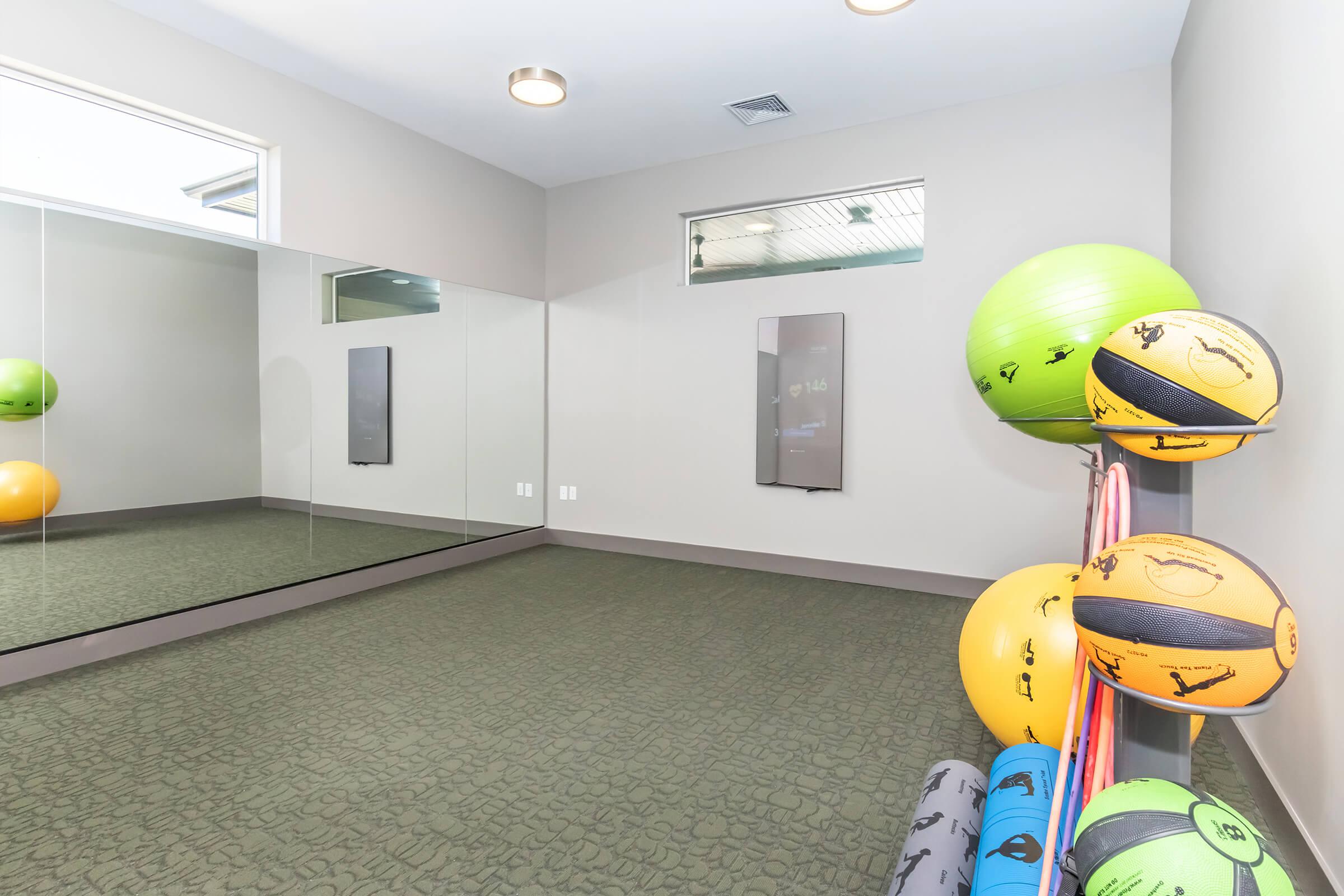 A bright fitness studio featuring a large mirror on one wall, with colorful exercise balls and mats stacked in the corner. The room has a neutral color palette and modern lighting, creating an inviting space for workouts.