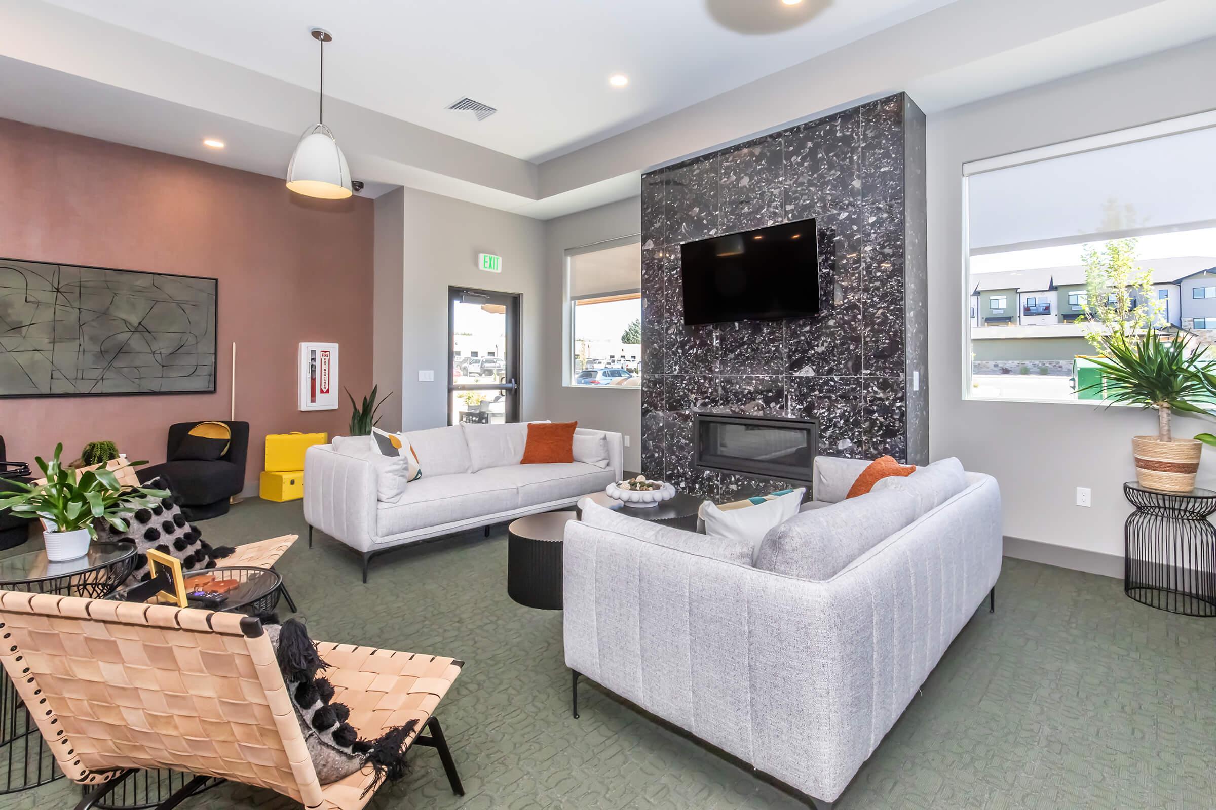 A modern living area featuring two gray sofas with orange and black accents, a sleek black stone wall with a mounted TV, and large windows allowing natural light. Potted plants and decorative items add a touch of greenery, while a unique chair and a small side table complete the stylish space.