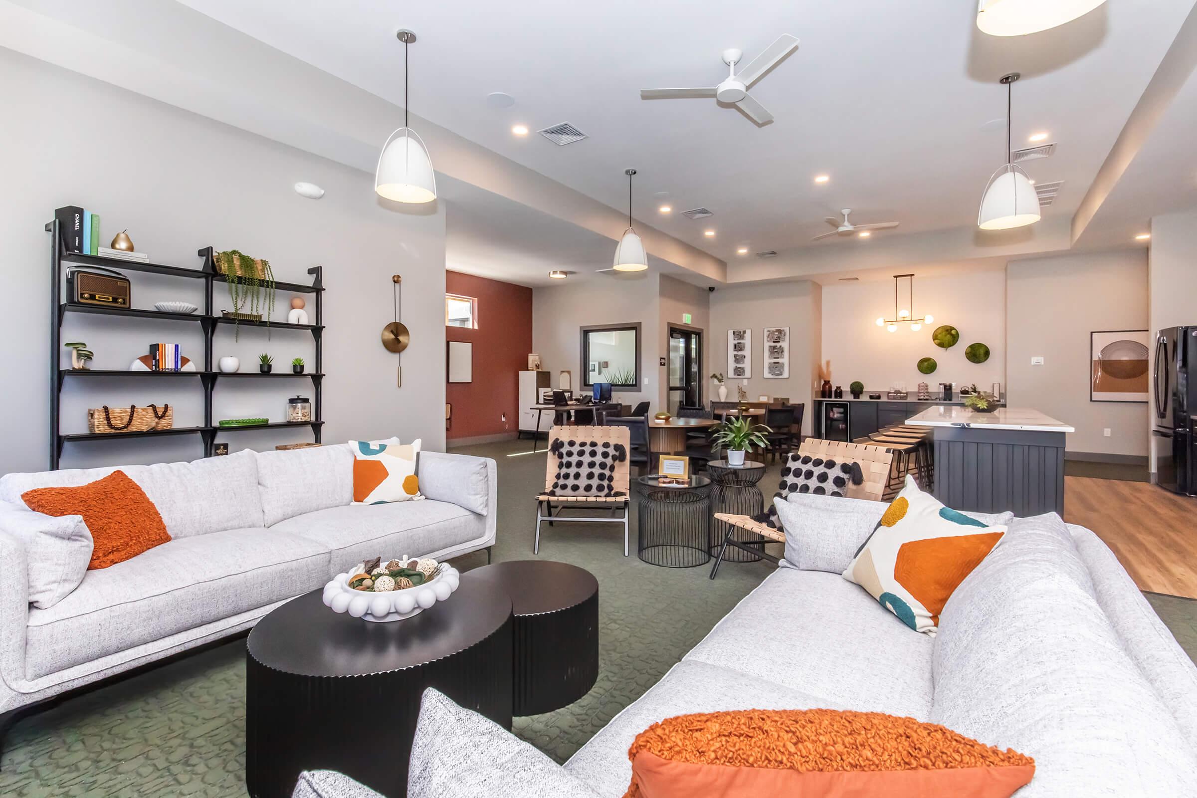 A stylish and modern living room featuring two light gray sofas with decorative pillows, a round coffee table, indoor plants, and a black shelving unit. The space is well-lit with ceiling fans and includes a dining area visible in the background. Neutral-toned walls enhance the contemporary aesthetic.
