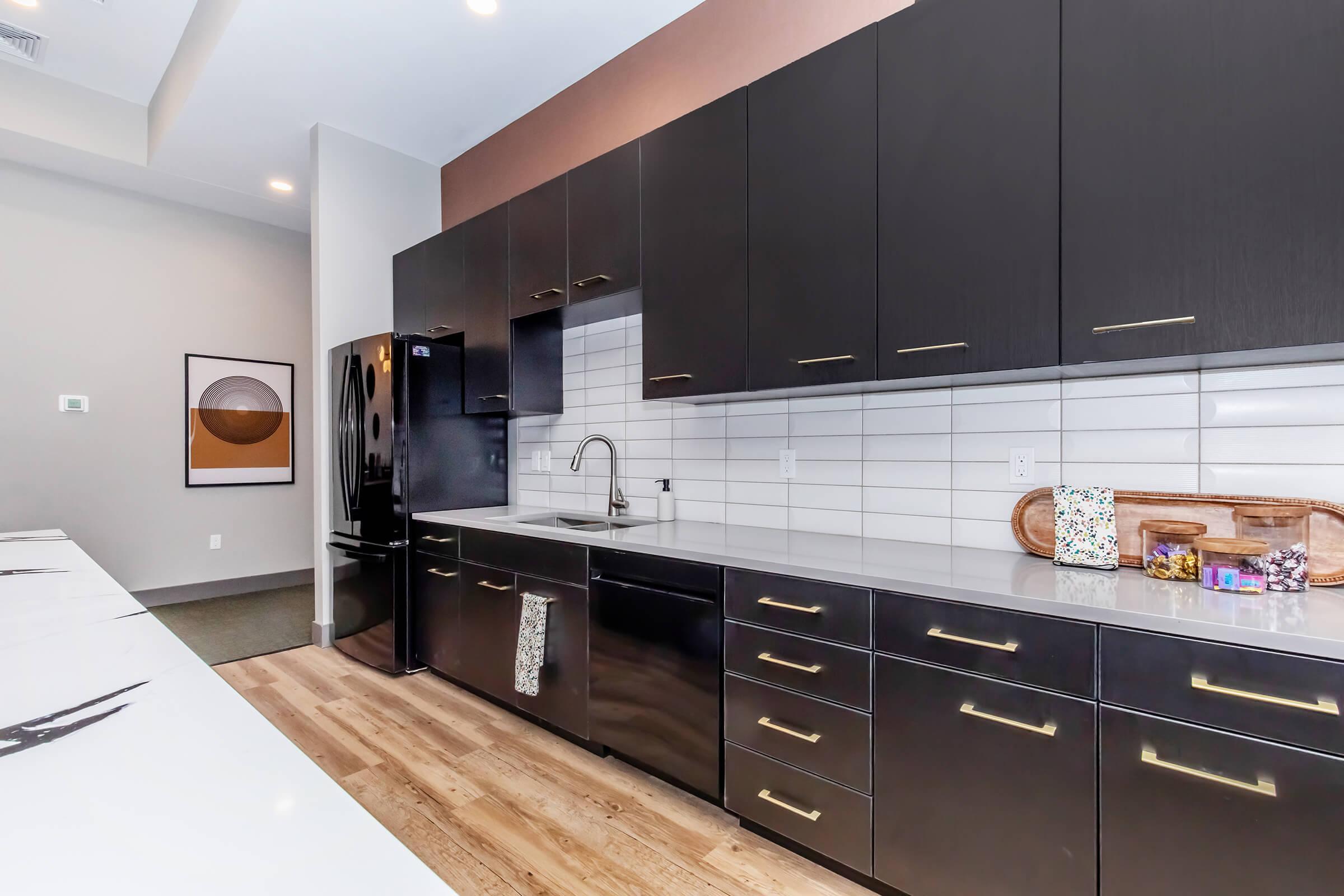Modern kitchen featuring sleek black cabinetry with gold hardware, a large sink, and a black refrigerator. The countertops are light-colored, complementing the cabinetry. A wooden cutting board and decorative items are arranged on the counter, and there's a stylish wall art piece in the background.