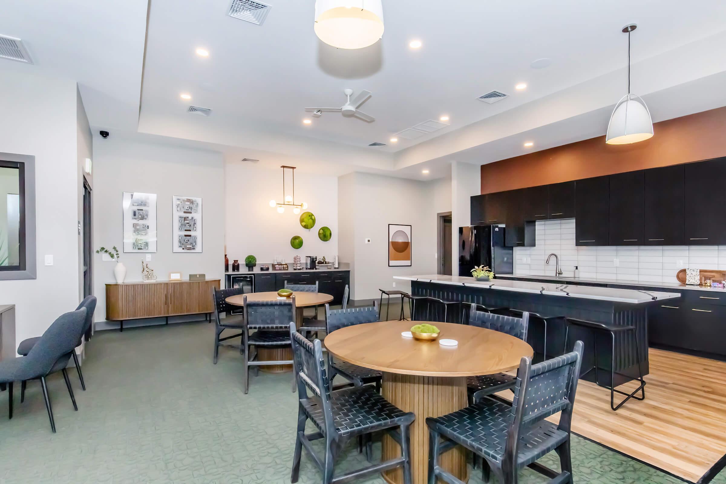 A modern community lounge interior featuring a round dining table with chairs, an island kitchen area with black cabinets, and a seating area. The space is well-lit with pendant lights, and decorative elements like wall art and green decor add a touch of style. A welcoming atmosphere for social gatherings.