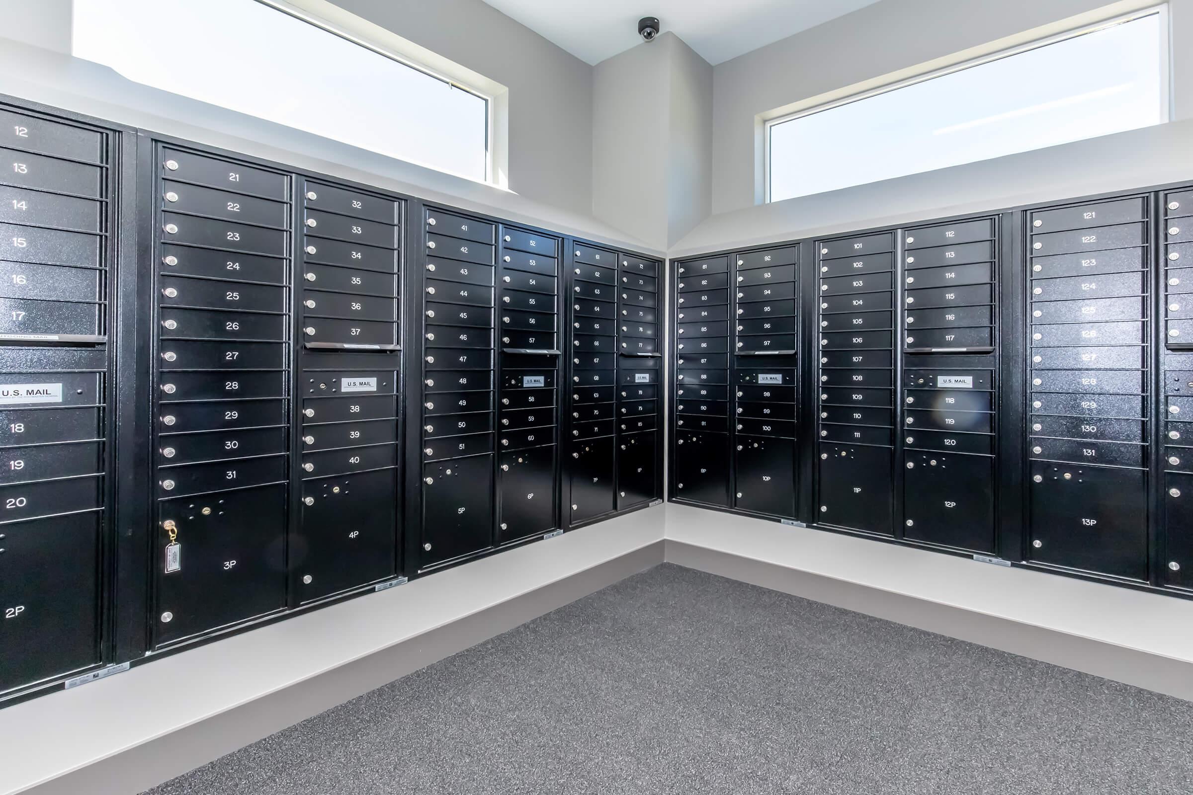 A secure postal room featuring multiple black mailbox units arranged against the walls. Each mailbox has a numbered slot and a small keyhole, with large windows allowing natural light to enter the space. The floor is covered with dark carpet, creating a clean, organized appearance.