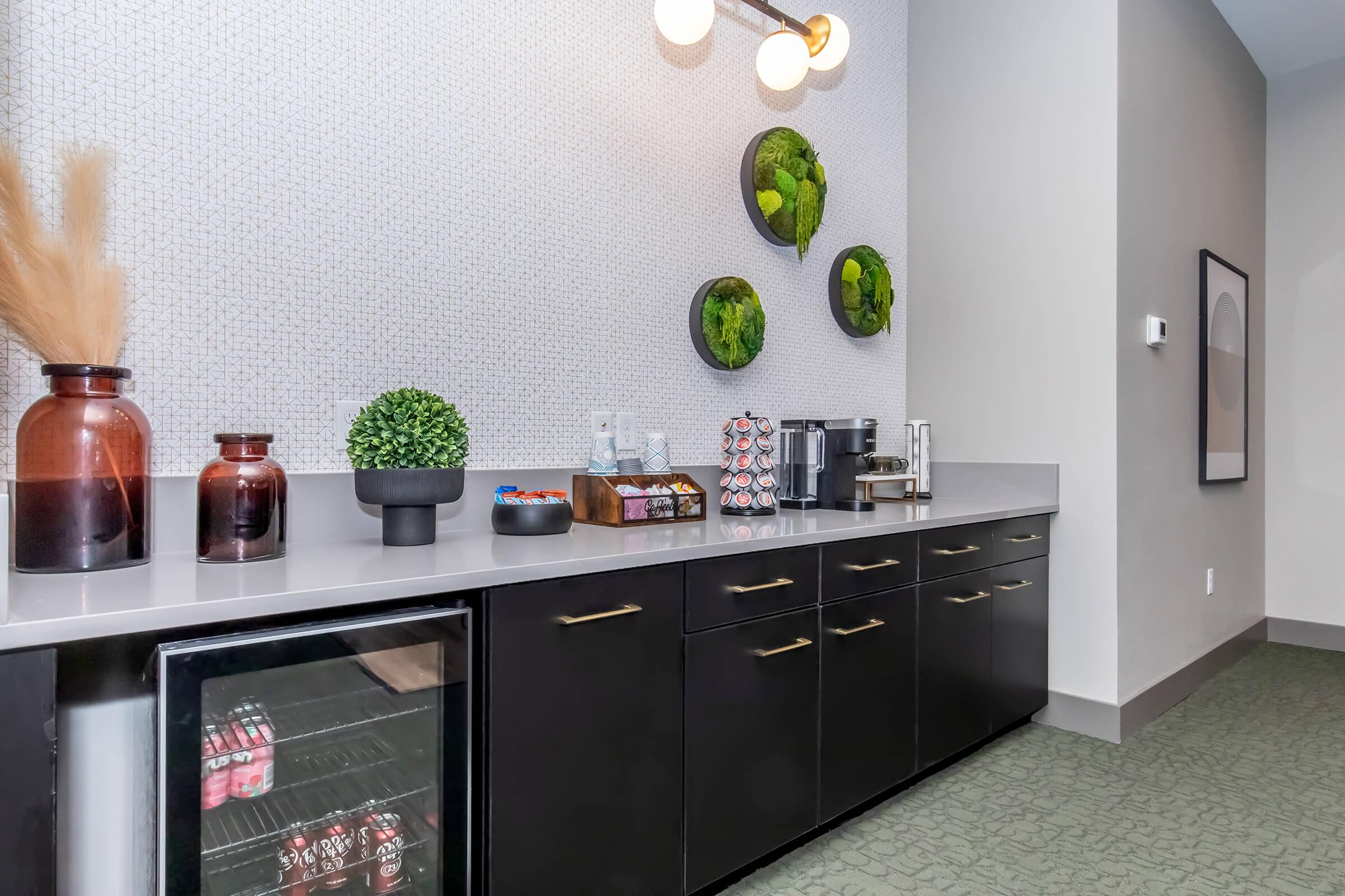 A modern kitchenette featuring a countertop with a variety of snacks, a coffee maker, and decorative plants on the wall. Below the countertop, there's a small refrigerator displaying drinks. The sleek cabinetry and stylish wall design add a contemporary touch to the space.