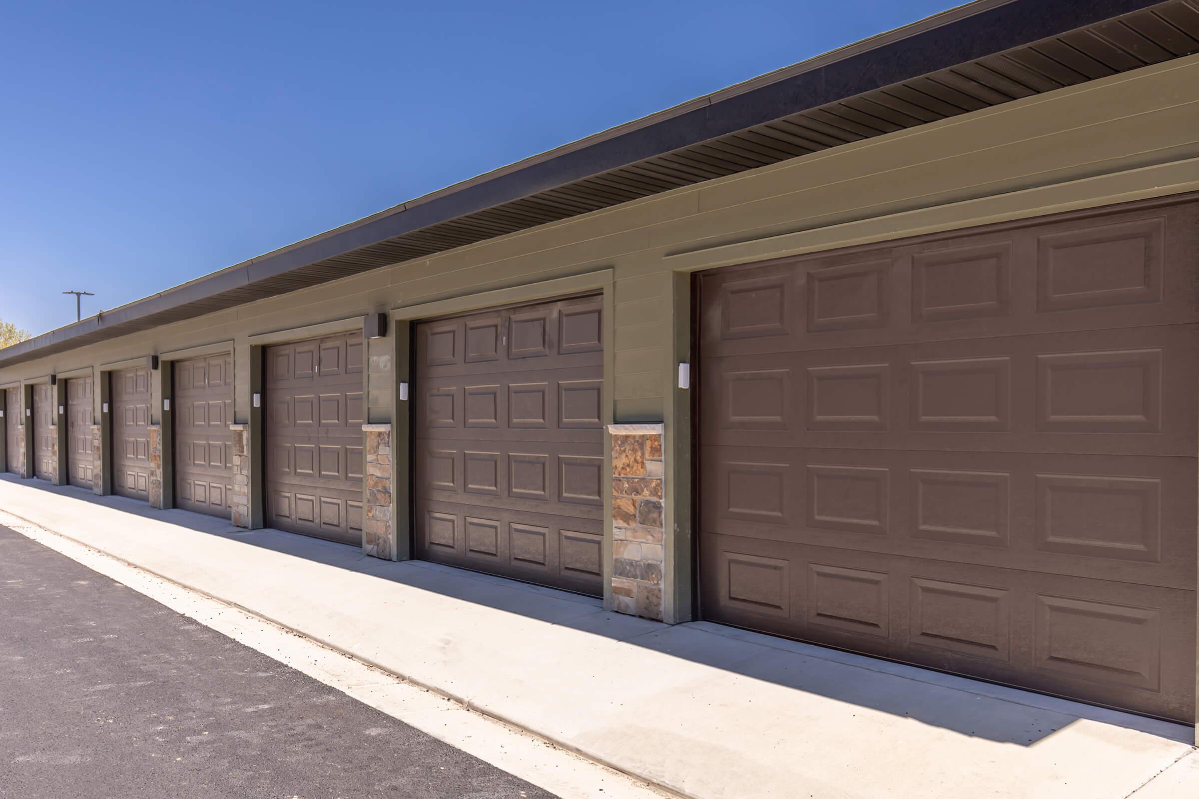 A row of closed storage unit garage doors with a concrete sidewalk in front. The buildings are painted in a neutral color with stone accents at the base. The clear blue sky is visible above, indicating a sunny day.