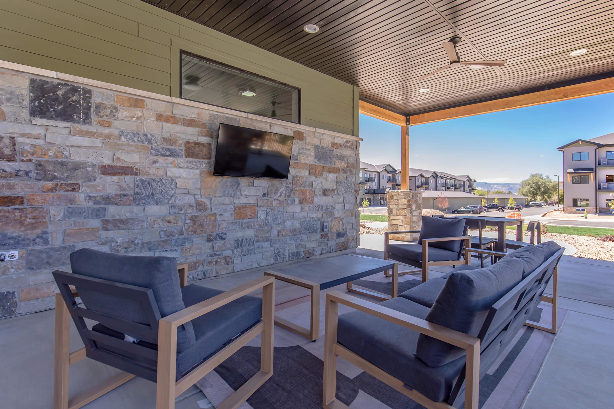 An outdoor lounge area featuring modern wooden chairs and tables set against a stone wall. A wall-mounted TV is visible. The space is partially shaded by a wooden overhang, and nearby residential buildings can be seen in the background, suggesting a community setting.