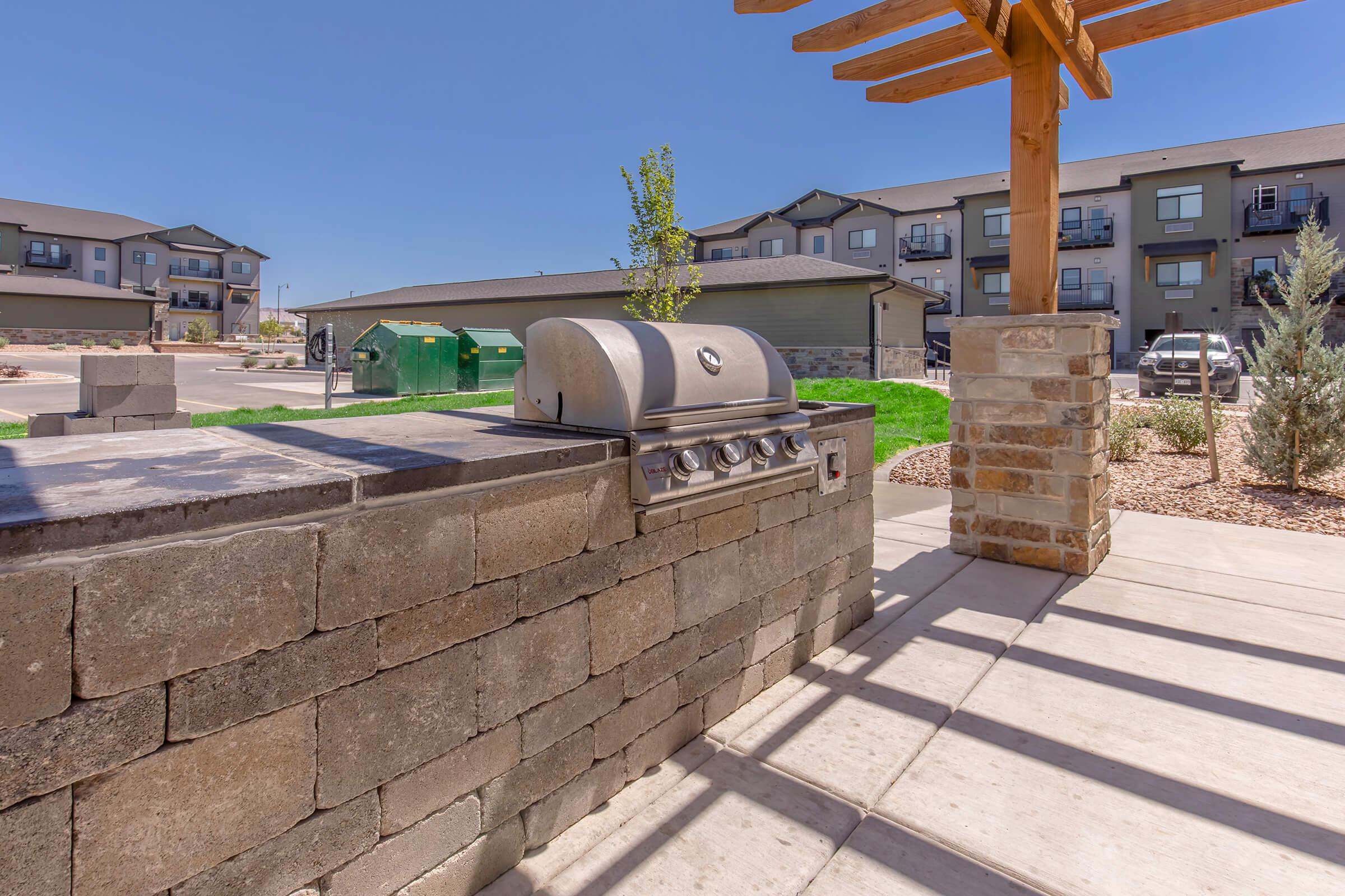 Outdoor grilling area featuring a stainless steel gas grill on a stone countertop, set in a landscaped environment with modern apartment buildings in the background and a wooden pergola casting shadows. Grass and gravel landscaping complement the space.