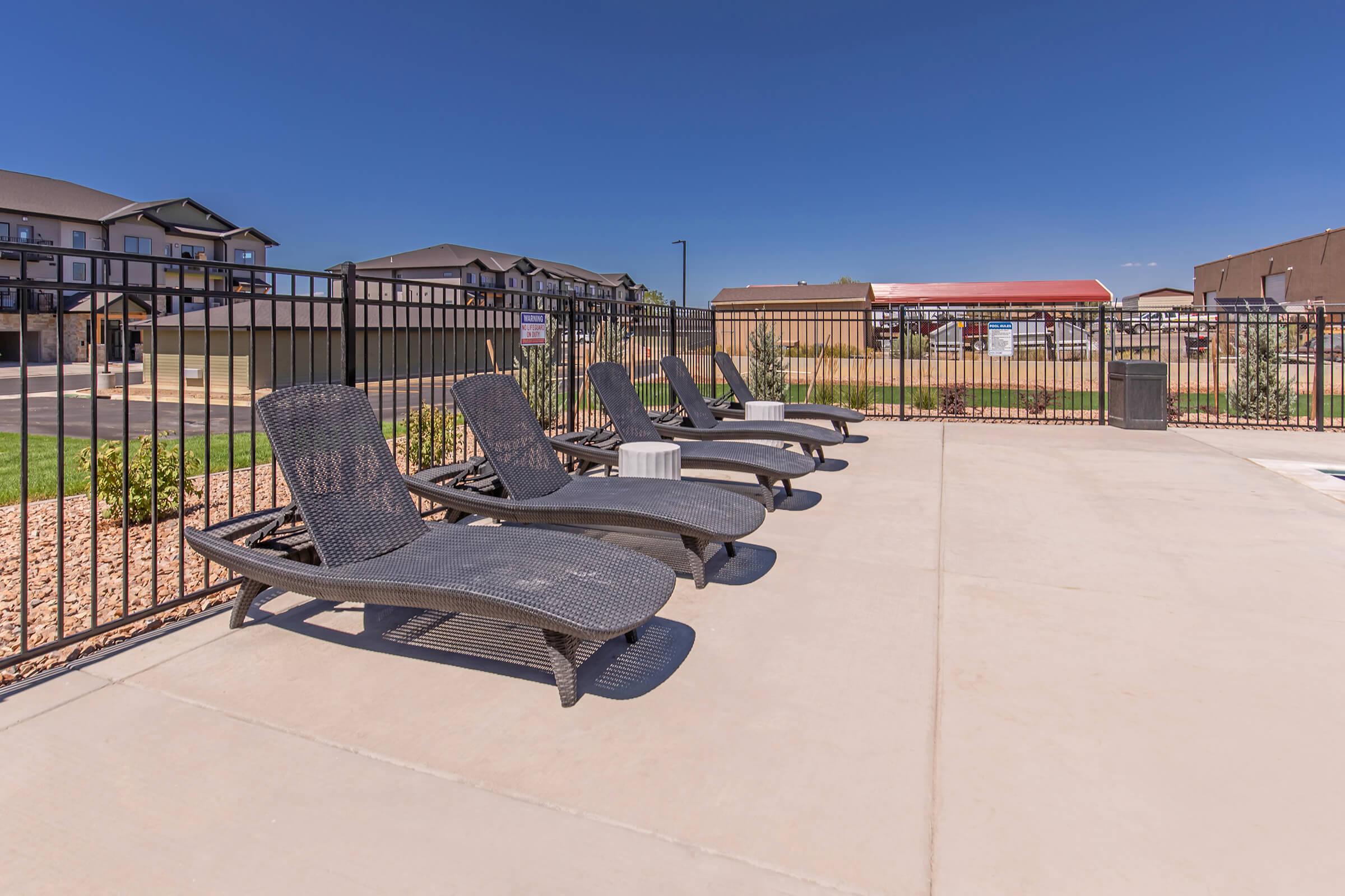 A row of black lounge chairs by a pool area, surrounded by a metal fence. The background features residential buildings and a clear blue sky. The concrete deck is clean and well-maintained, with a landscaped area visible.