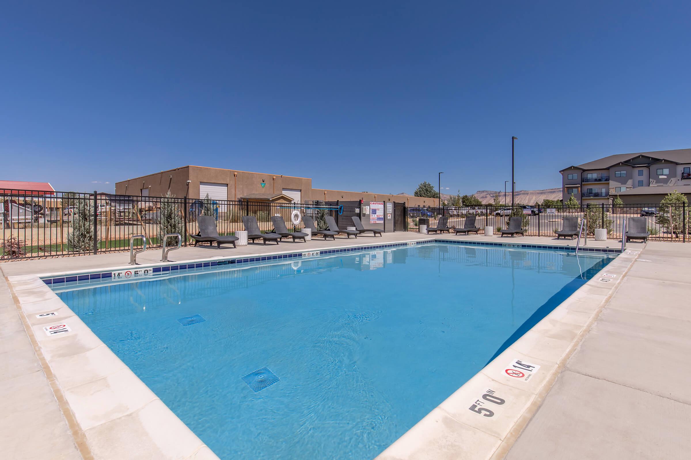A clear blue swimming pool surrounded by lounge chairs and a safety fence. In the background, there are buildings and a clear sky. The pool area invites relaxation and leisure activities under the bright sun.