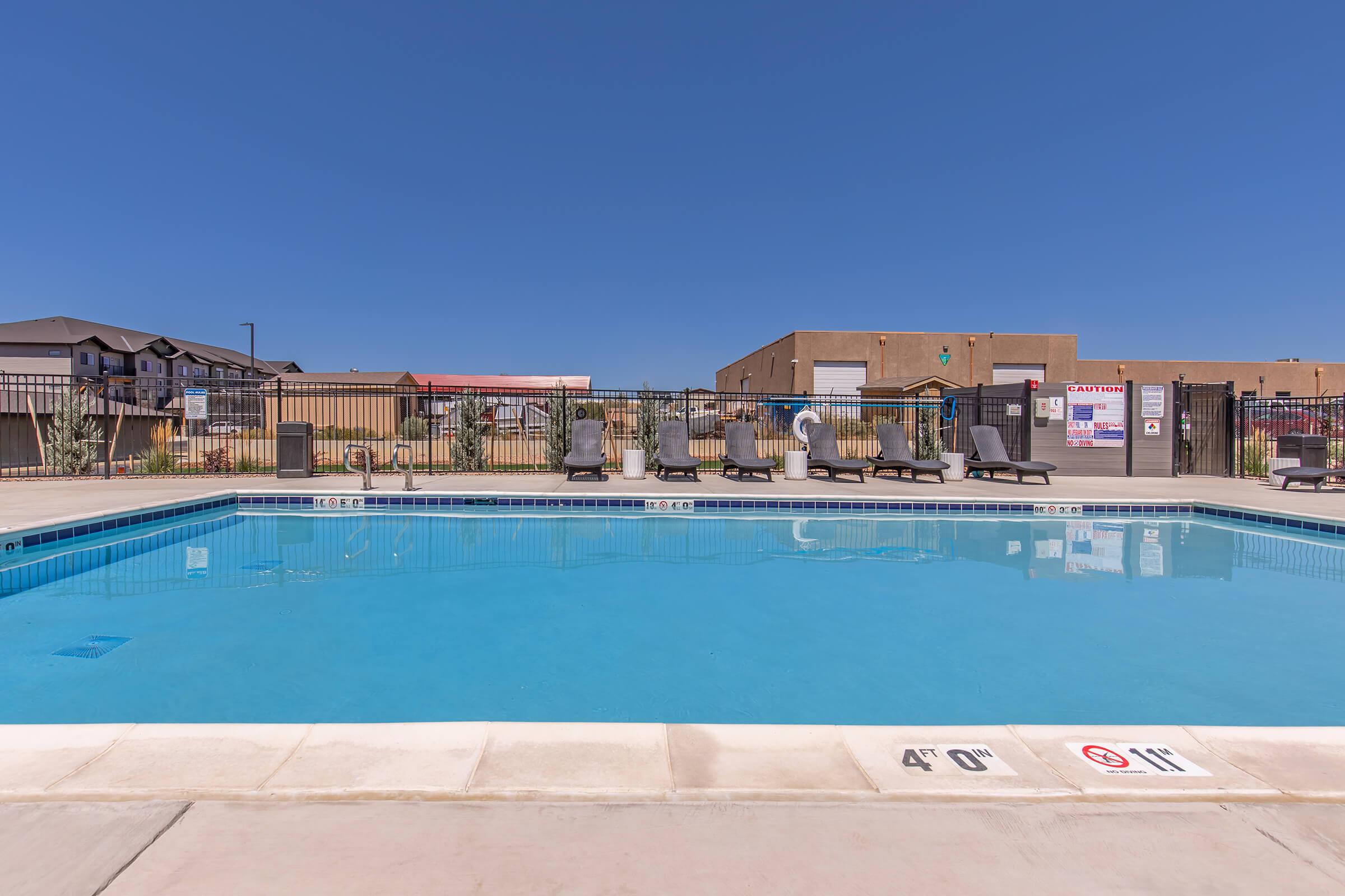 A clear, inviting swimming pool surrounded by lounge chairs. The pool features a depth marker showing 4 feet. In the background, there are buildings and a fence, with a bright blue sky overhead. The scene is well-lit and suggests a leisure area suitable for relaxation and enjoyment.