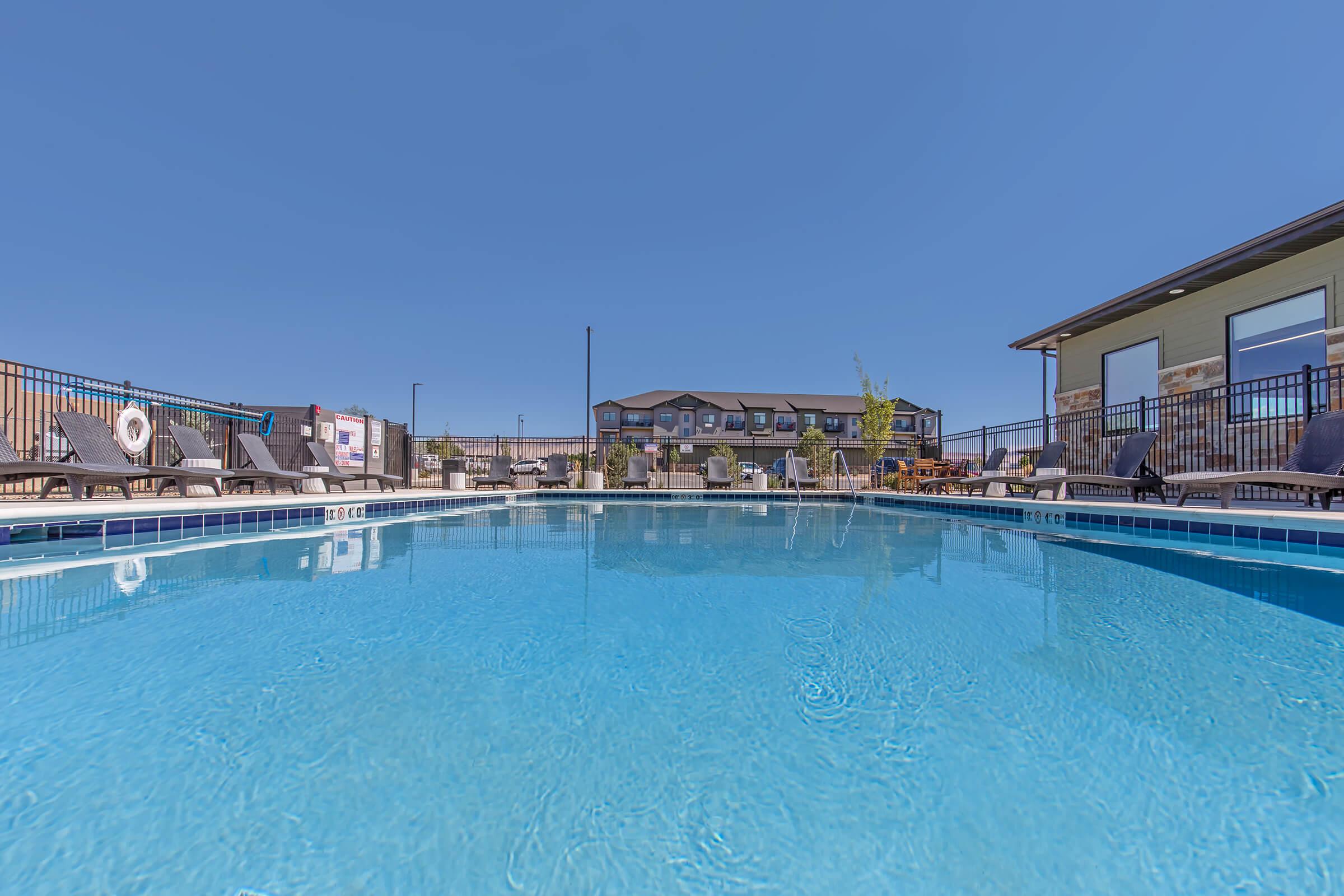 A clear, inviting swimming pool on a sunny day, with lounge chairs around the pool's edge. In the background, there are buildings and a clear blue sky, creating a relaxing atmosphere for outdoor leisure.