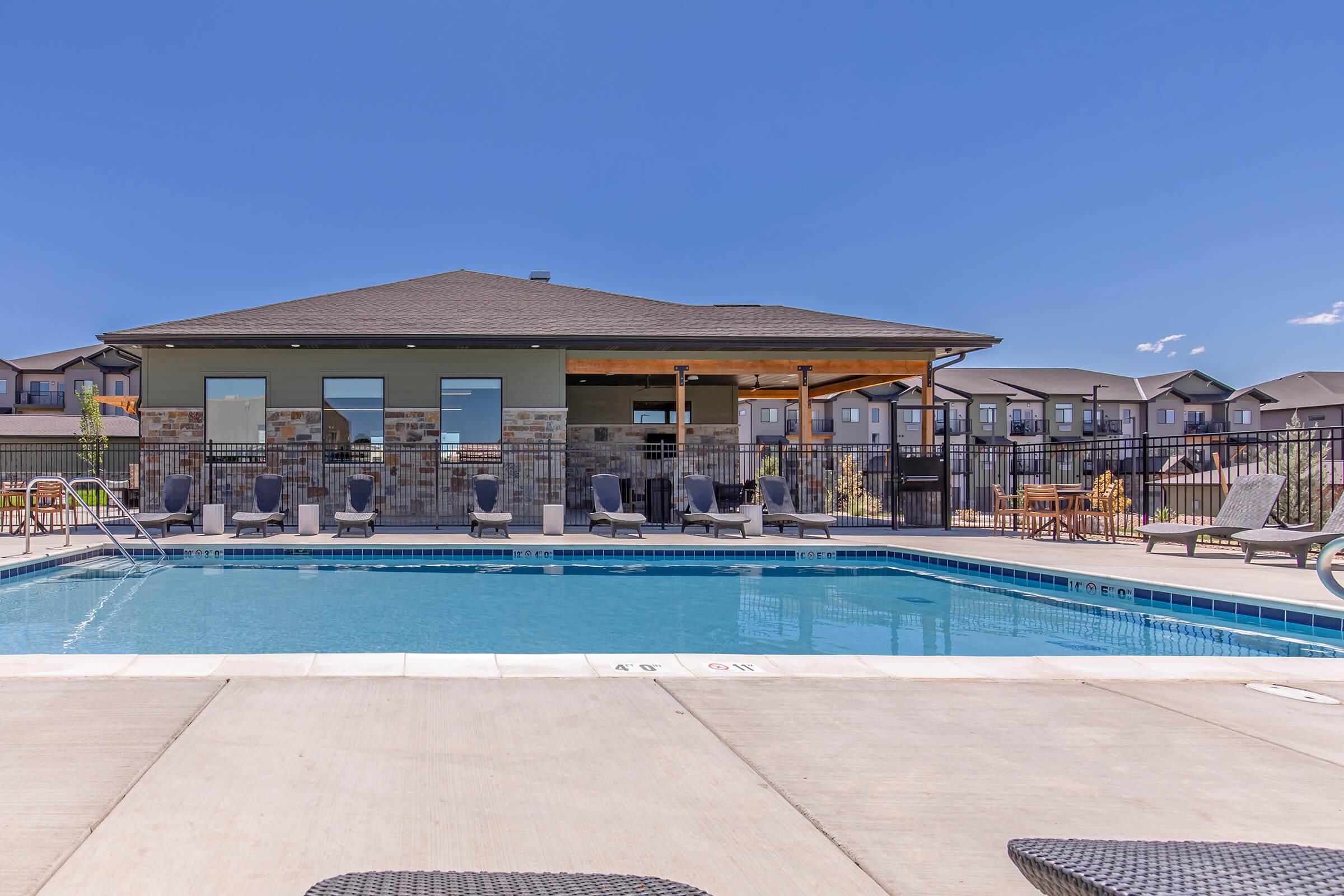 A serene outdoor swimming pool surrounded by a patio. There are several lounge chairs positioned around the pool, and a modern building with large windows can be seen in the background. The sky is clear and blue, creating a relaxing atmosphere for leisure activities.
