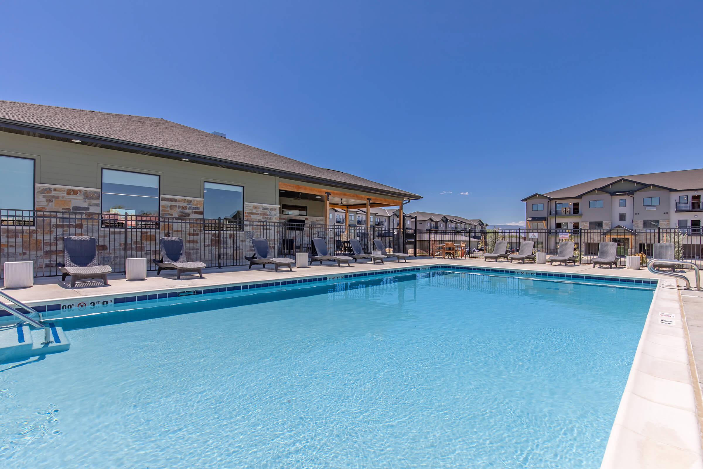 A clear swimming pool surrounded by lounge chairs and a fence, with a modern building visible in the background. The sky is bright blue, creating a sunny atmosphere. The scene conveys a relaxing outdoor space, ideal for leisure and enjoyment.