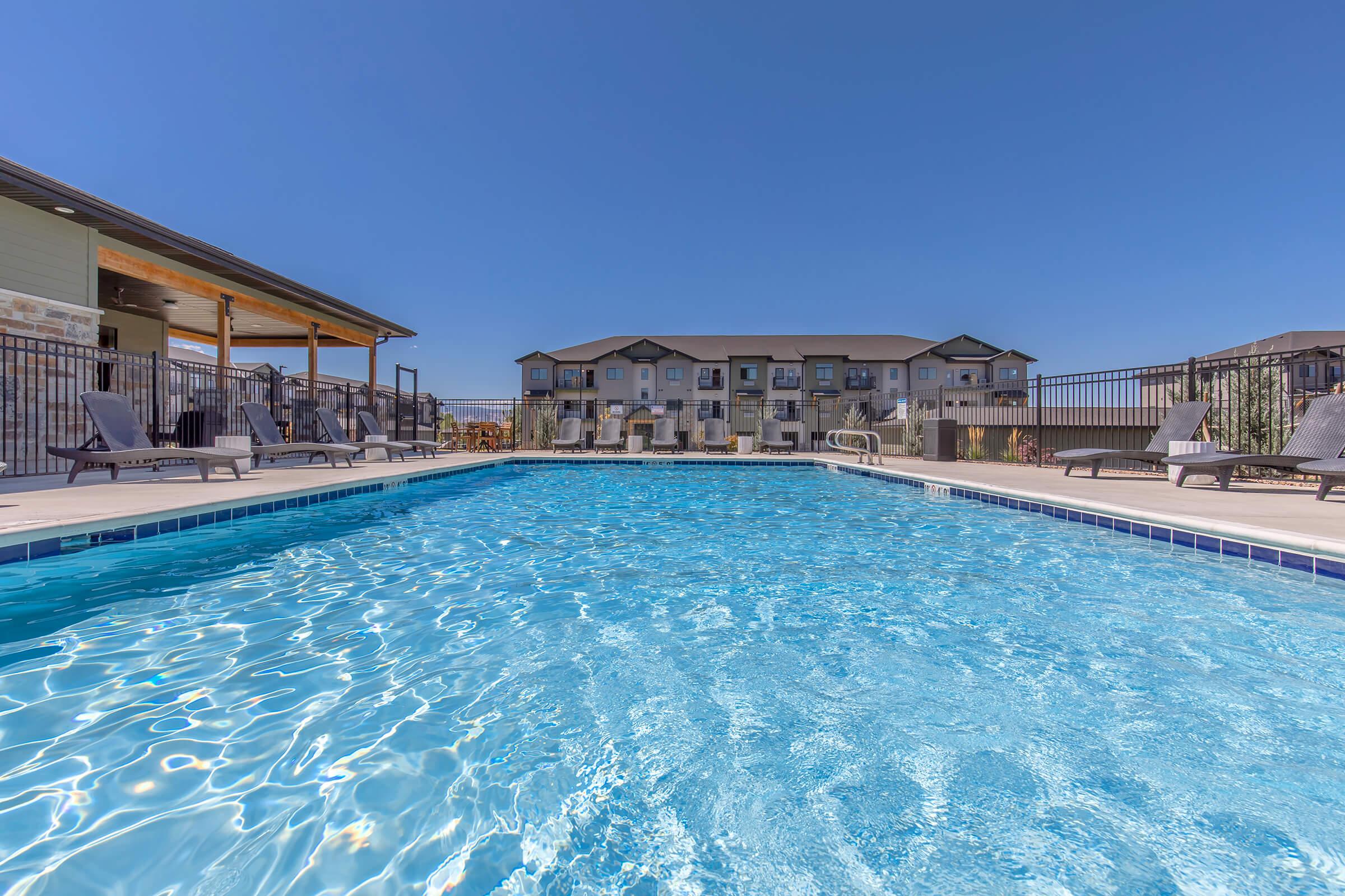 A clear blue swimming pool with lounge chairs around it, set against a bright blue sky. In the background, a multi-story building is visible, partially shaded by the poolside structure. The scene suggests a relaxing outdoor leisure area, ideal for recreation and unwinding.