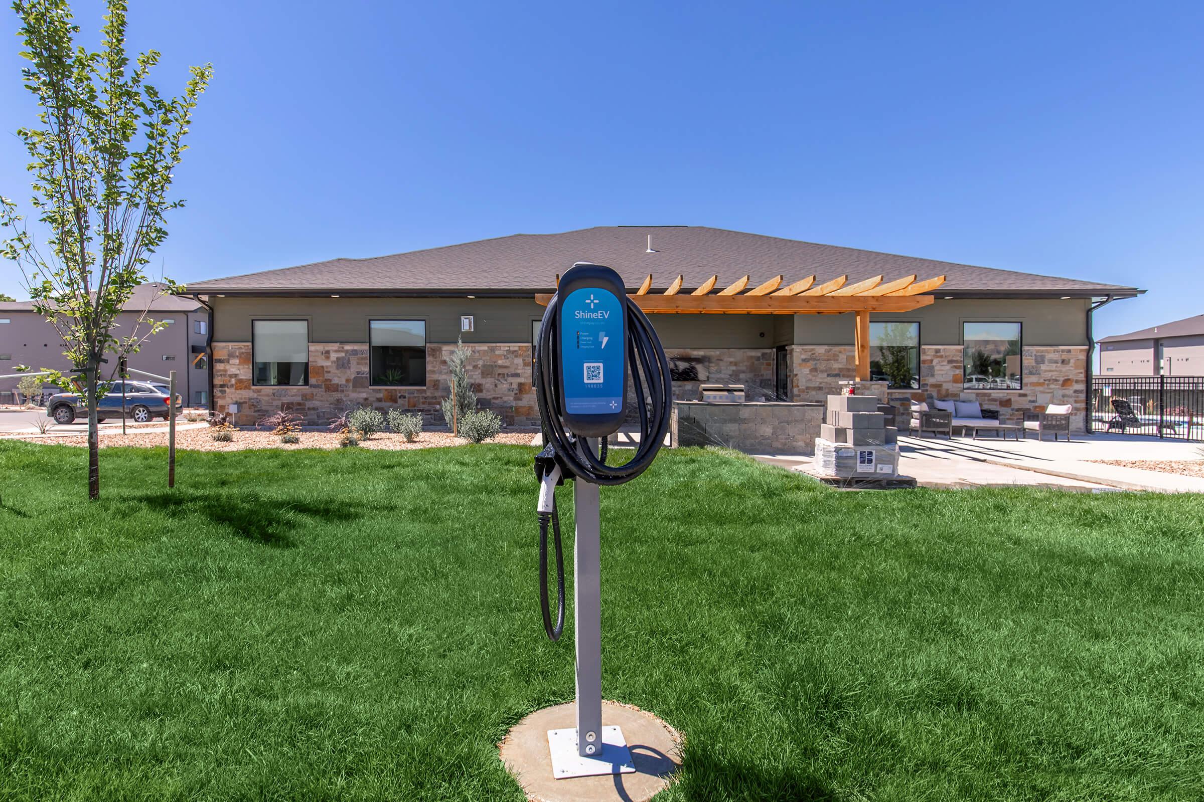 An electric vehicle charging station stands prominently in a grassy area, with a modern building featuring a stone facade and a covered patio in the background. Clear blue skies and neatly landscaped surroundings enhance the inviting atmosphere.