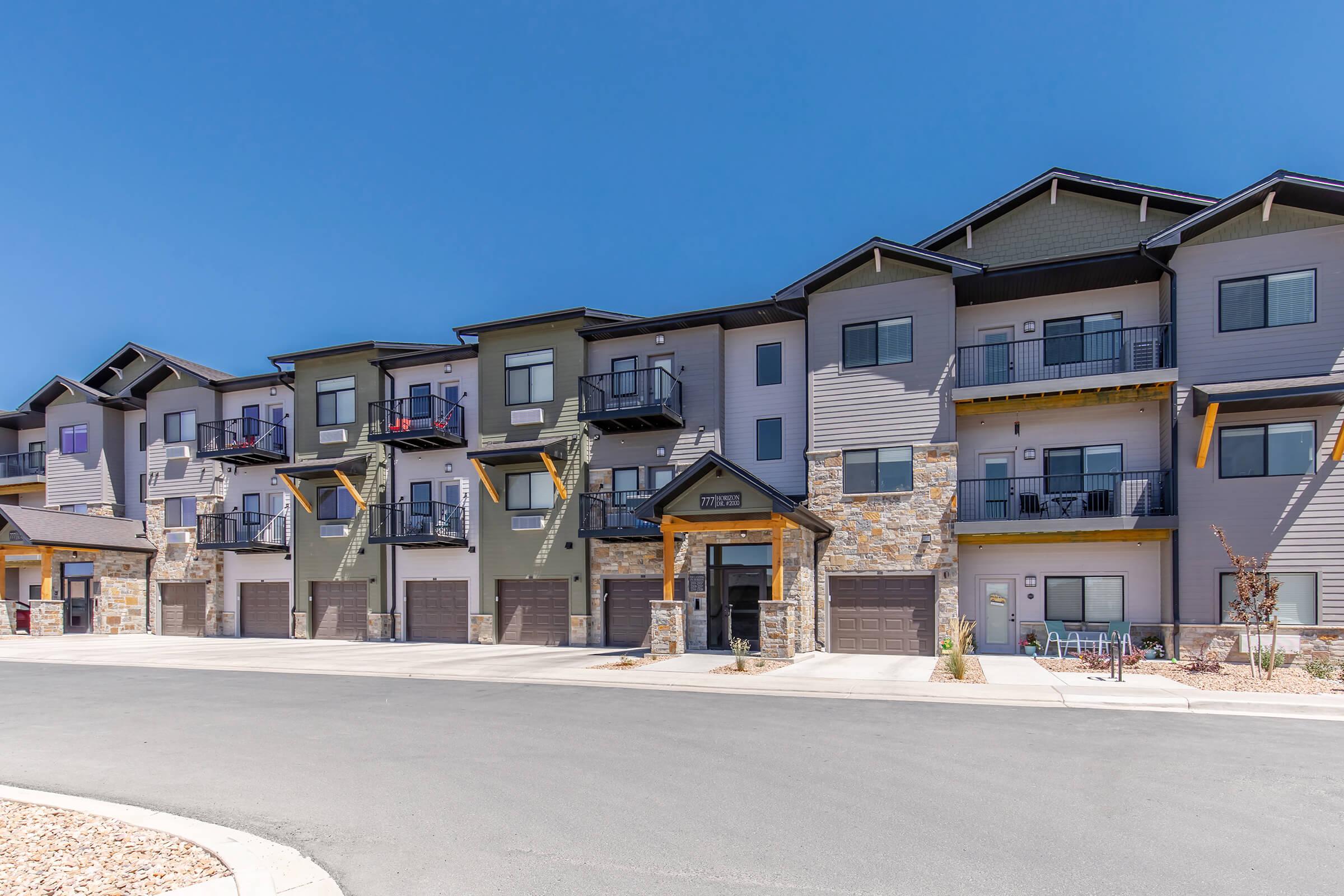 A modern multi-unit apartment building featuring a blend of stone and siding exteriors. The structure has multiple balconies, garage spaces, and a welcoming entrance under a wooden canopy. The surrounding area is paved with a clear blue sky overhead.