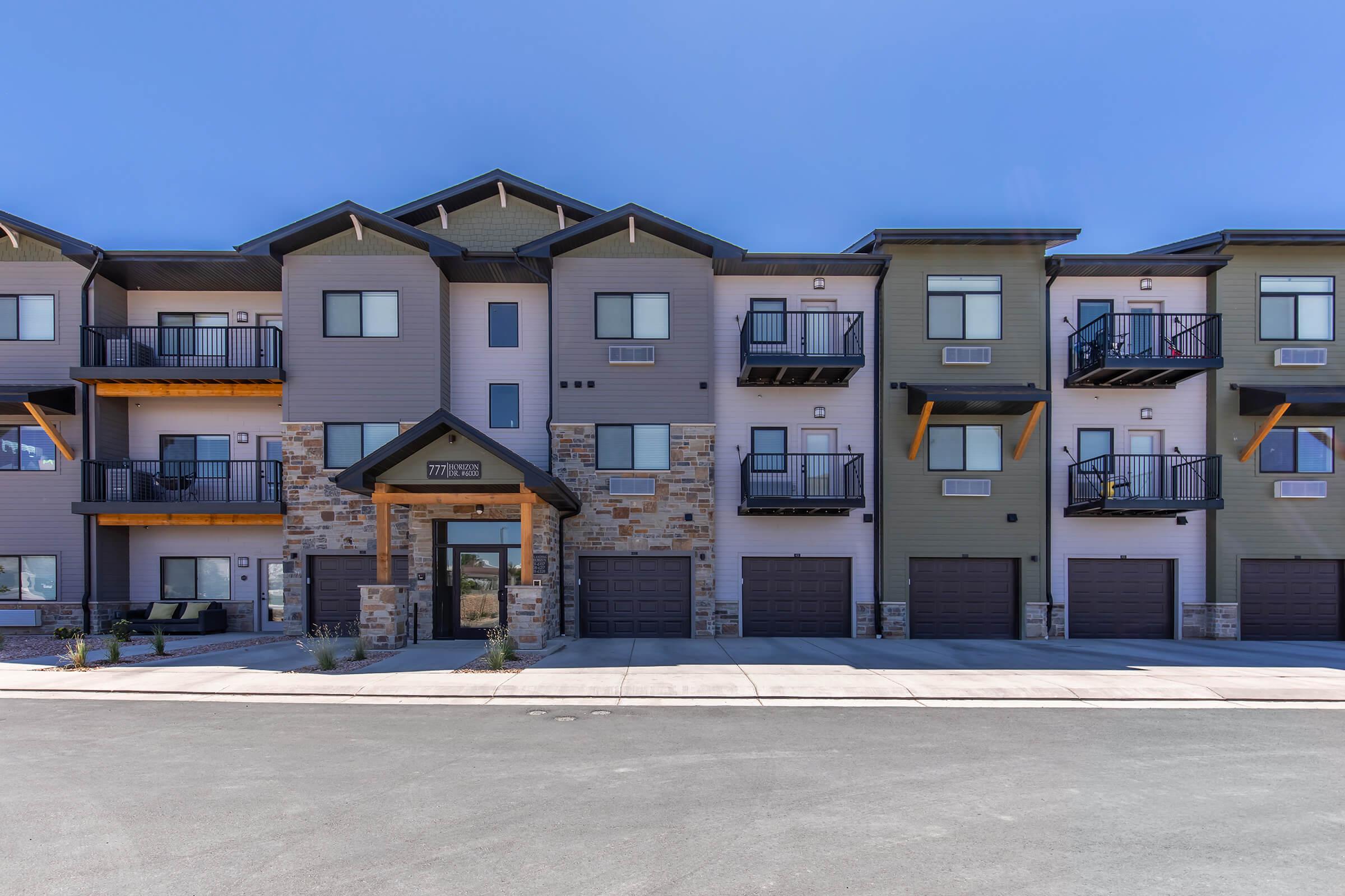 A modern apartment building featuring multiple units with balconies, stone accents, and attached garages. The structure has a contemporary design with a combination of light and dark colors, set against a clear blue sky, surrounded by a paved area.