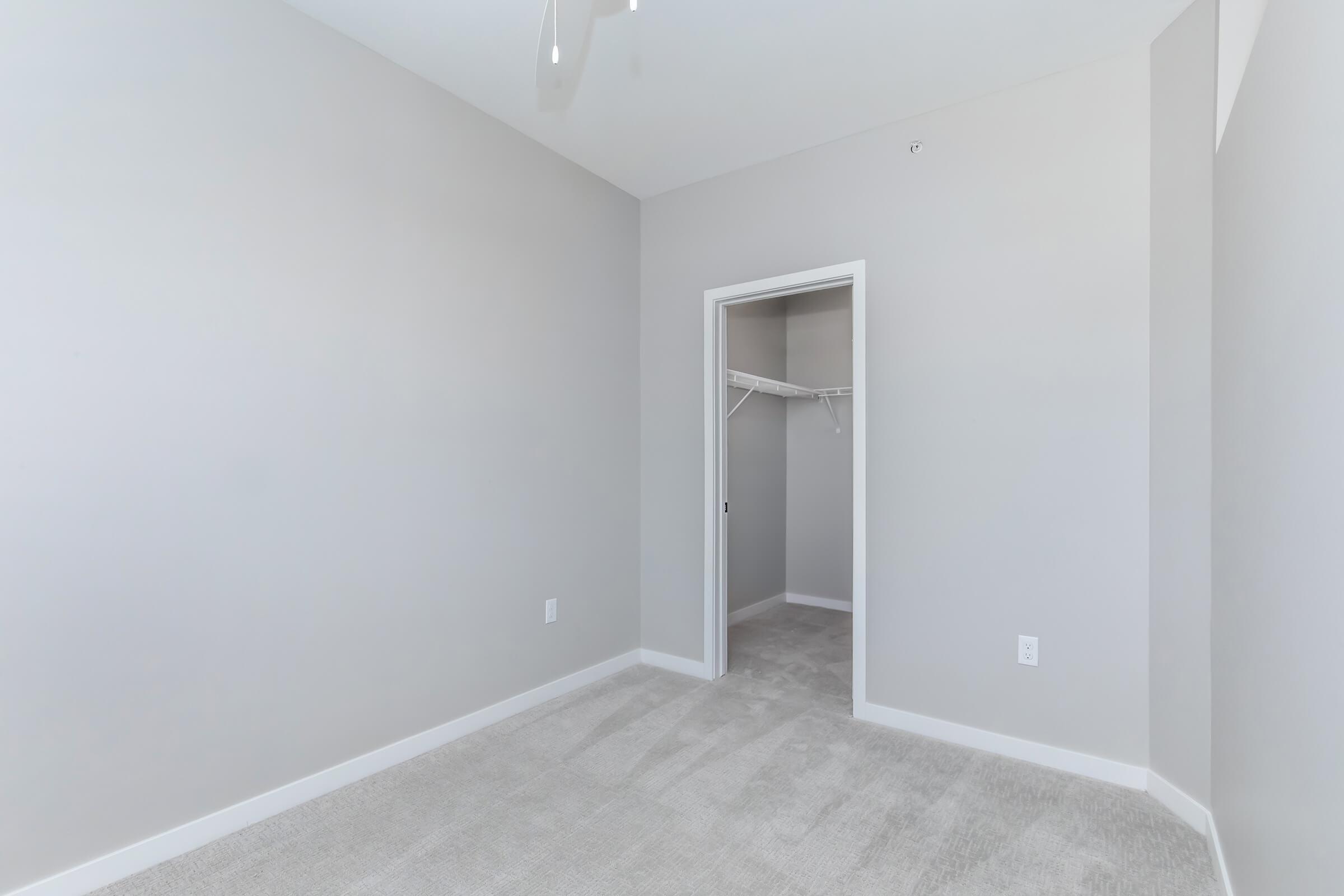 Empty room with light gray walls and carpeted floor. A doorway leads to a closet with a hanging rod. The ceiling features a ceiling fan, and there are no furnishings or decorations in the space, creating a minimalist and spacious appearance.