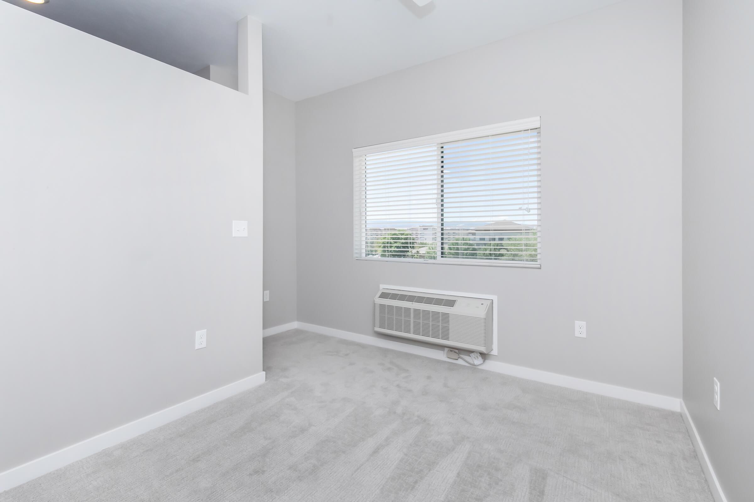 A small, light gray room with carpeted flooring and a window featuring blinds. An air conditioning unit is installed below the window. The walls are also light gray, creating a minimalistic and modern aesthetic.