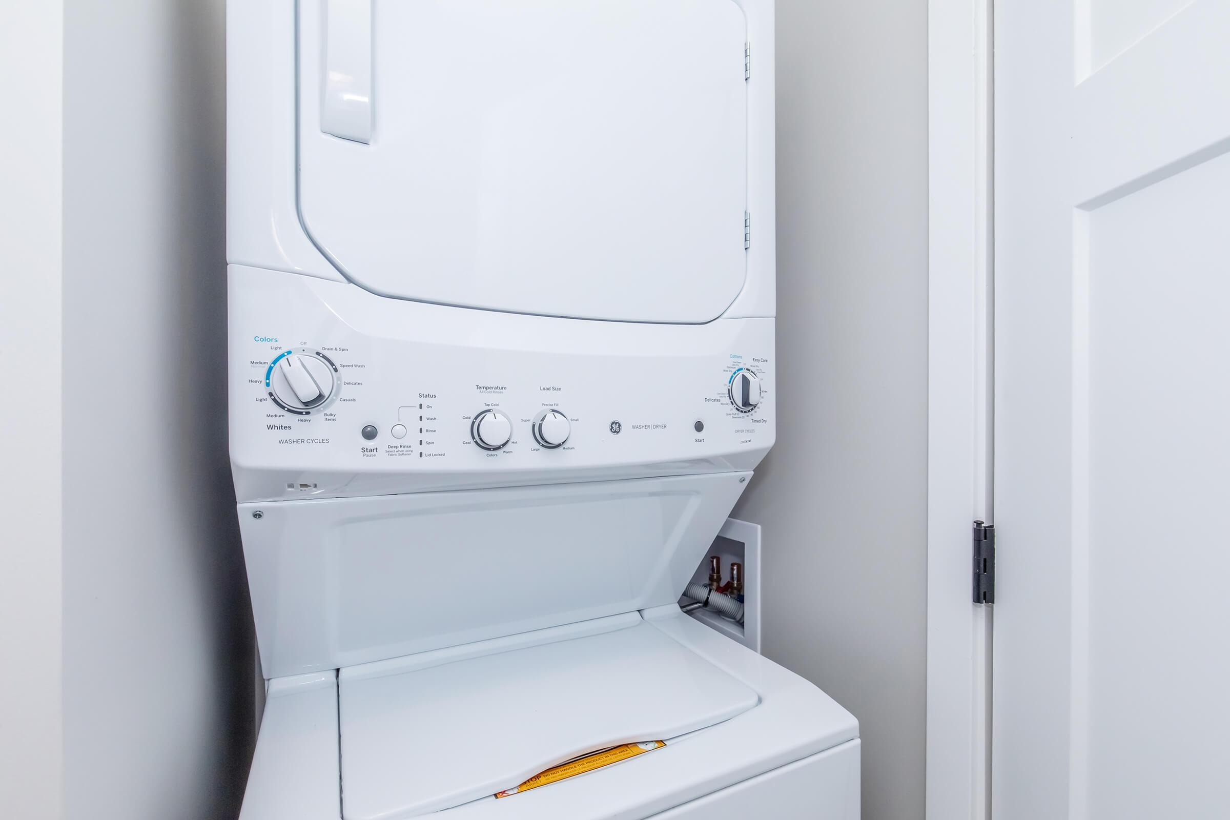 A white stacked washer and dryer unit located in a narrow laundry space. The controls for the machines are visible on the front, featuring various settings and knobs. The background shows a light-colored wall and a closed door.