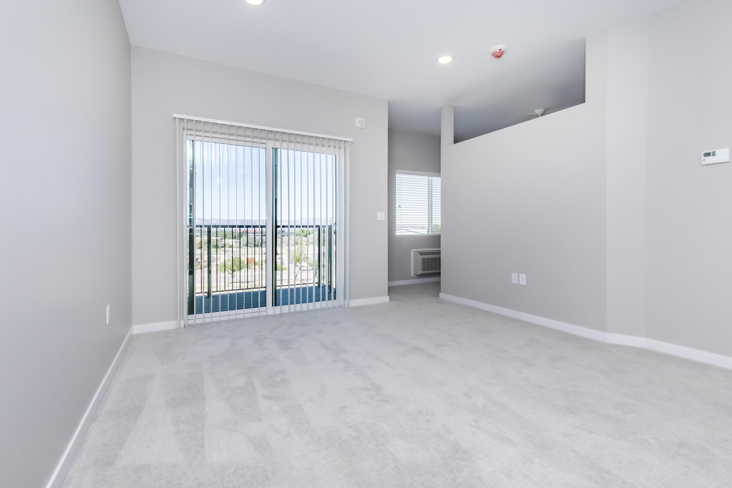 A modern, unfurnished living area with light gray walls and carpet. It features a large sliding glass door that opens to a balcony, providing natural light. A small window on the side allows additional light in. The space is open and airy, ideal for decorating and personalizing.