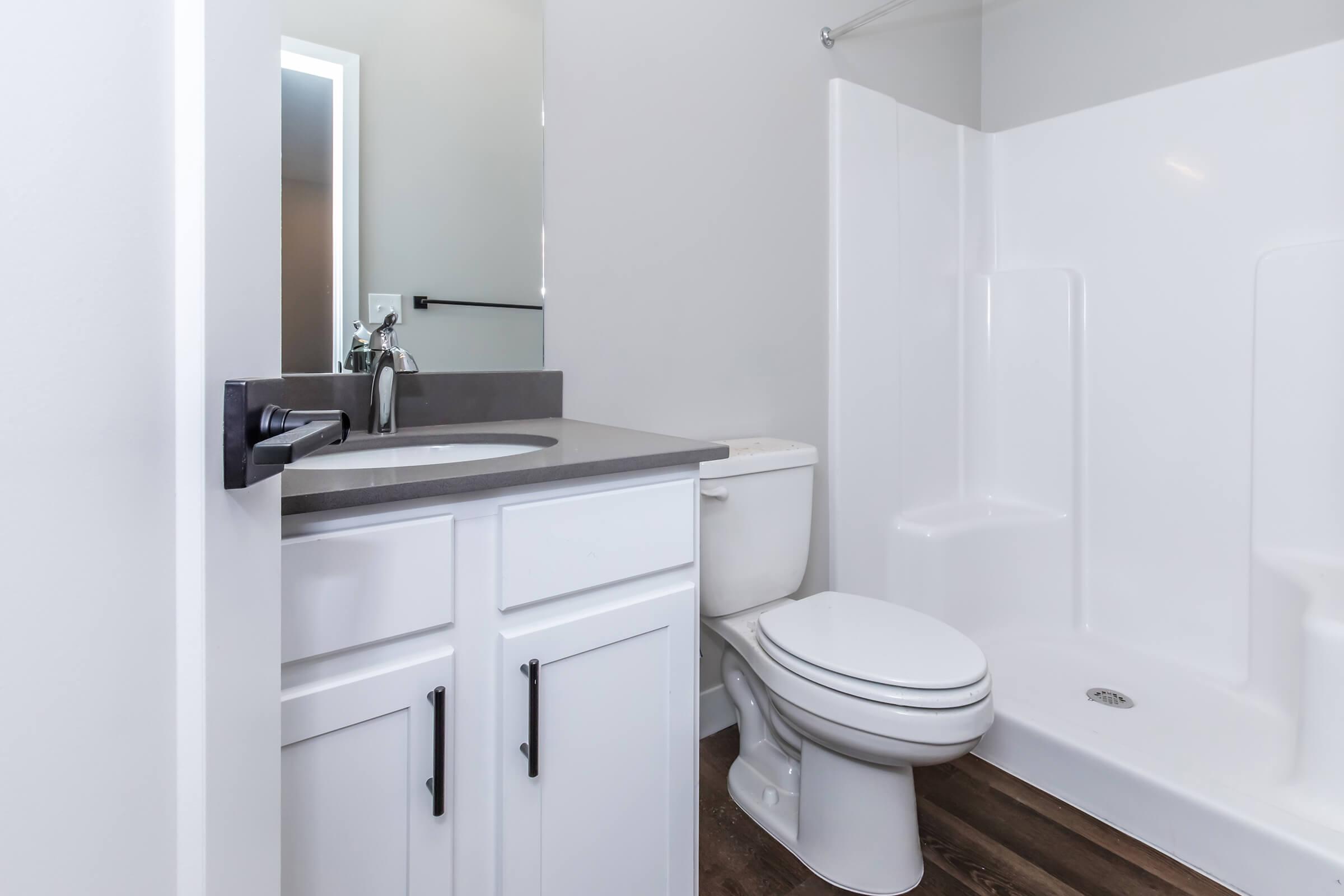 A clean, modern bathroom featuring a white shower/tub combination, a toilet, and a vanity with a sink. The walls are painted light gray, and there is a large mirror above the sink. The flooring is dark wood, adding contrast to the overall bright and spacious feel of the room.