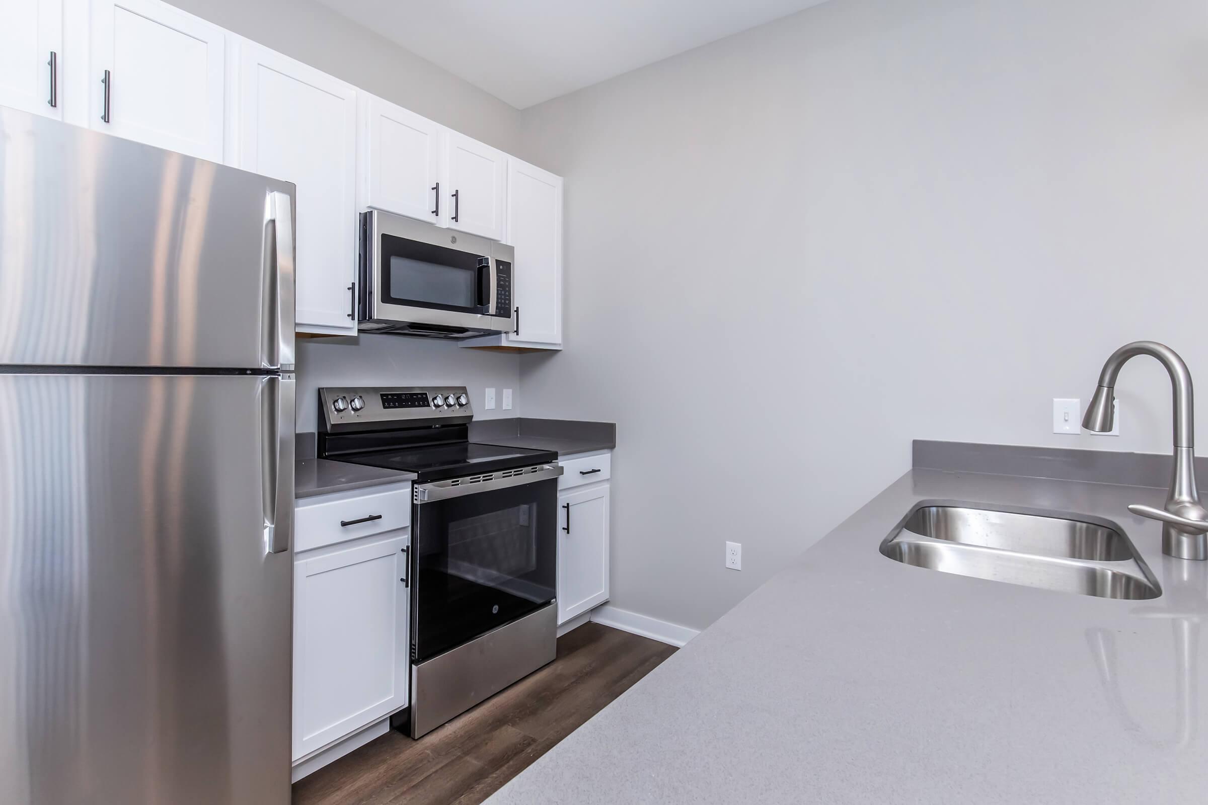 Modern kitchen featuring stainless steel appliances, including a refrigerator, microwave, and oven. The cabinetry is white with a sleek gray countertop and a stainless steel sink. The flooring is a warm wood tone, creating a clean and contemporary look. Walls are painted in a light gray color.