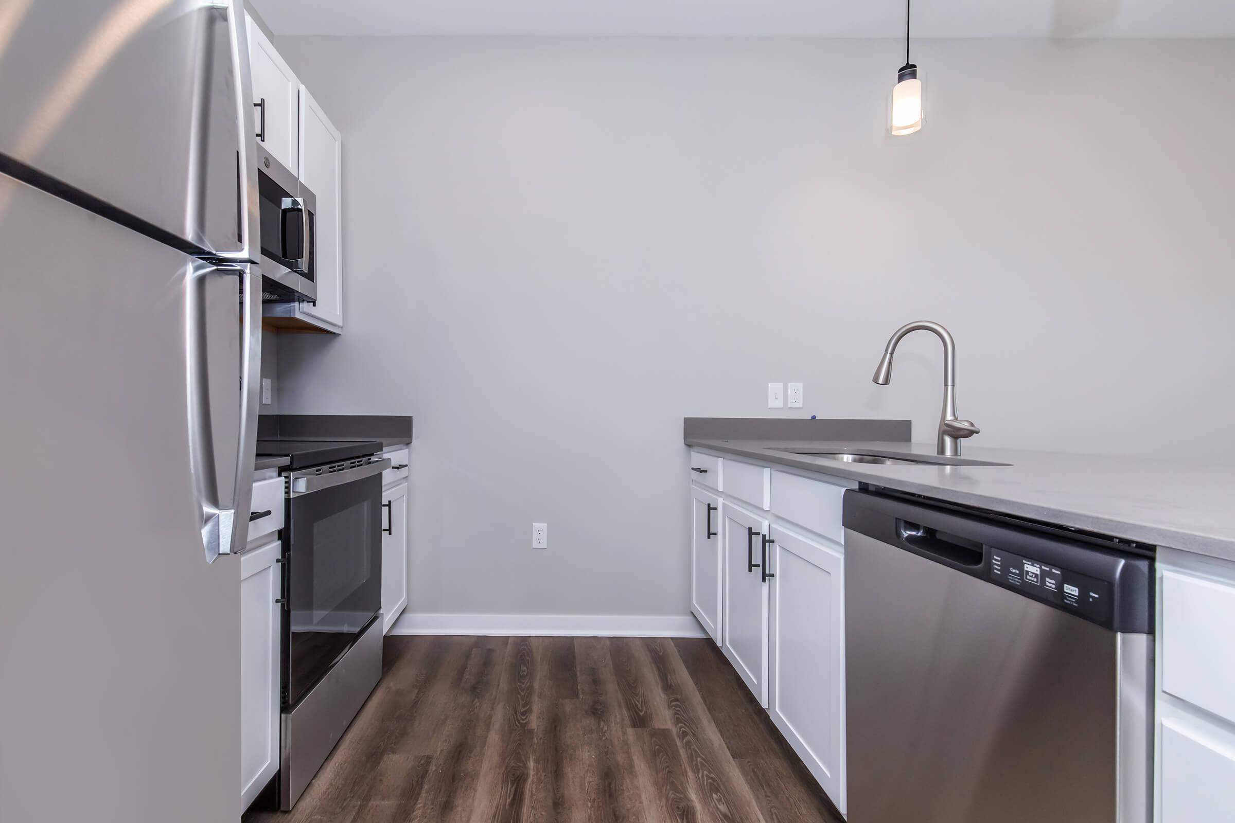 Modern kitchen featuring stainless steel appliances, including a refrigerator, oven, and dishwasher. The countertops are gray with a single sink and a stylish faucet. White cabinetry contrasts with the wood-like flooring, and natural light is provided by a pendant light hanging from the ceiling.