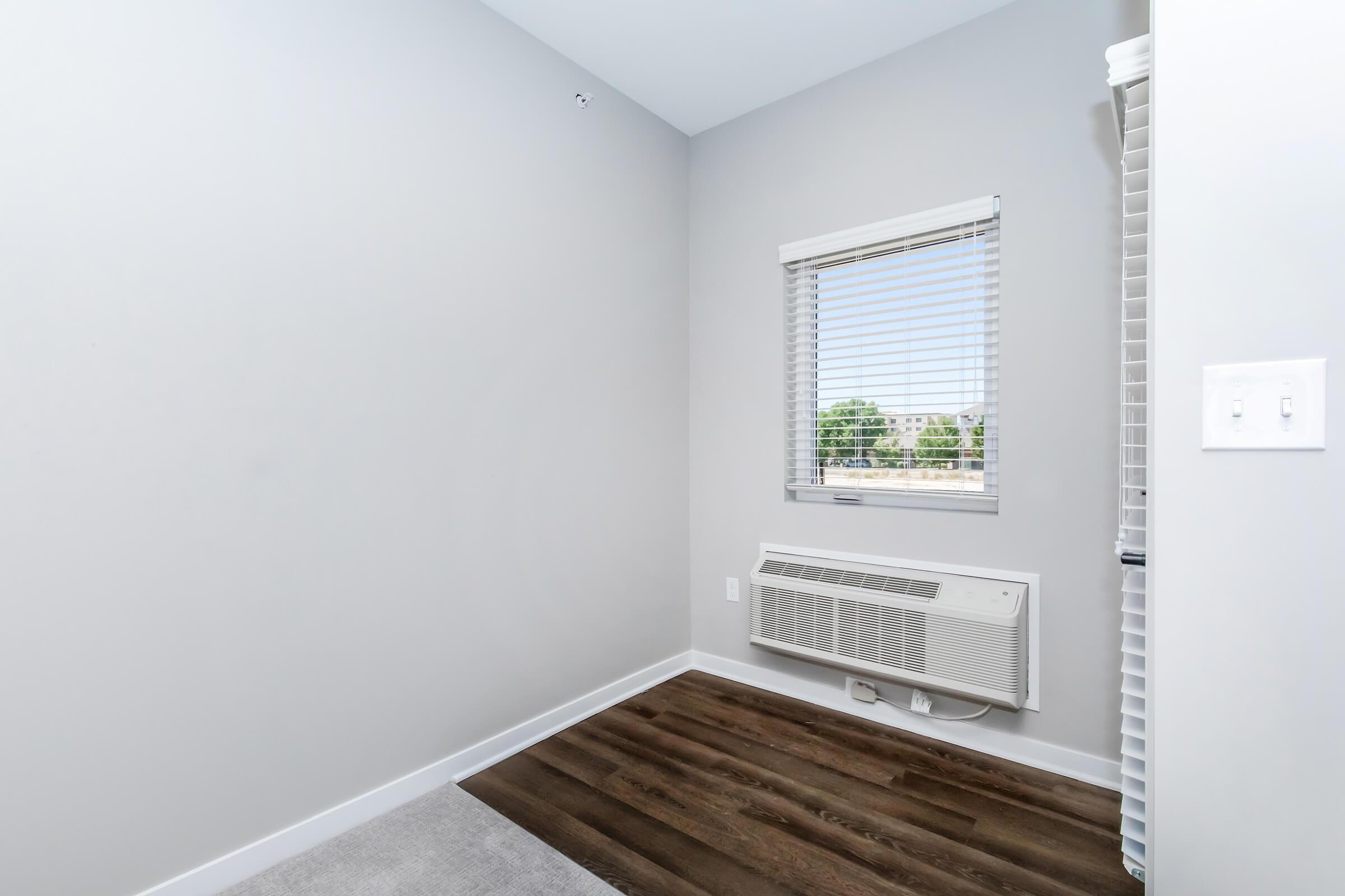 A minimalist interior corner of a room featuring light gray walls, a window with white blinds, a wall-mounted air conditioning unit, and a mix of wood and carpet flooring. Natural light enters through the window, illuminating the space.
