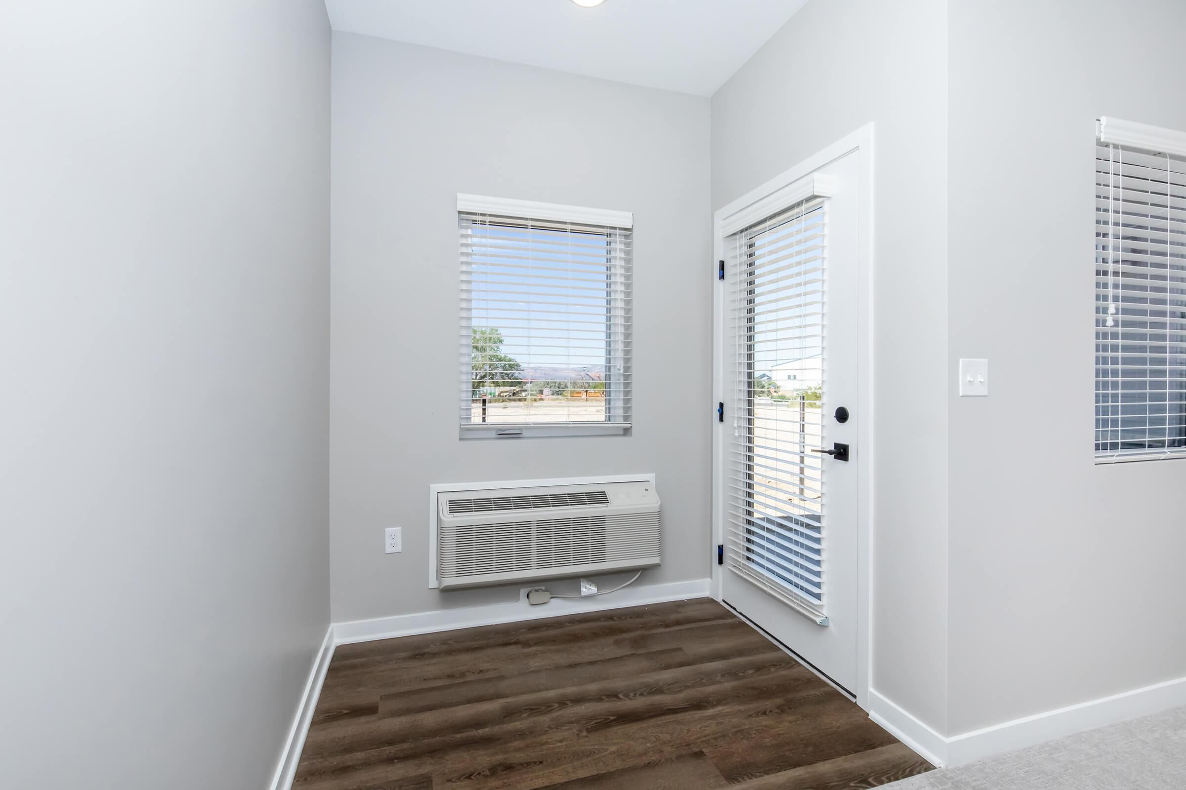 A clean, bright entryway featuring a door with a glass panel leading outside, a small window with blinds, and an air conditioning unit mounted on the wall. The floor is a light-colored wood laminate, and the walls are painted in a soft gray tone, creating a modern and welcoming atmosphere.