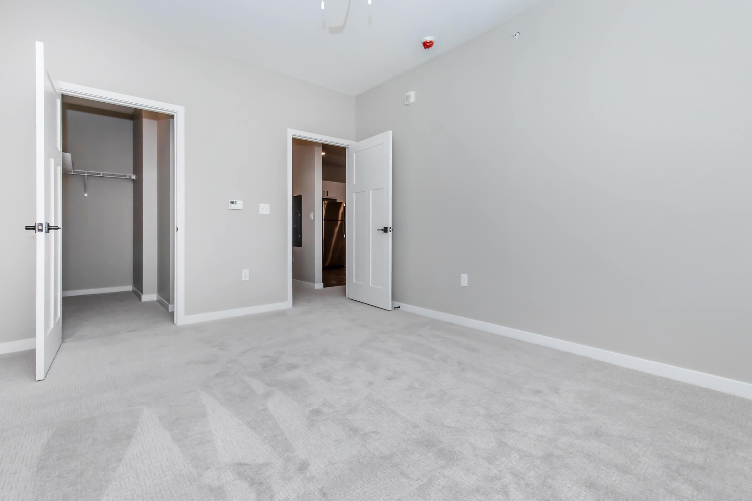 A spacious, empty room with light gray walls and plush beige carpeting. Two doors are open, leading to a closet and an adjacent space. Soft, natural light fills the room, highlighting its modern design and clean lines. The ceiling features a ceiling fan, adding to the room's airy feel.