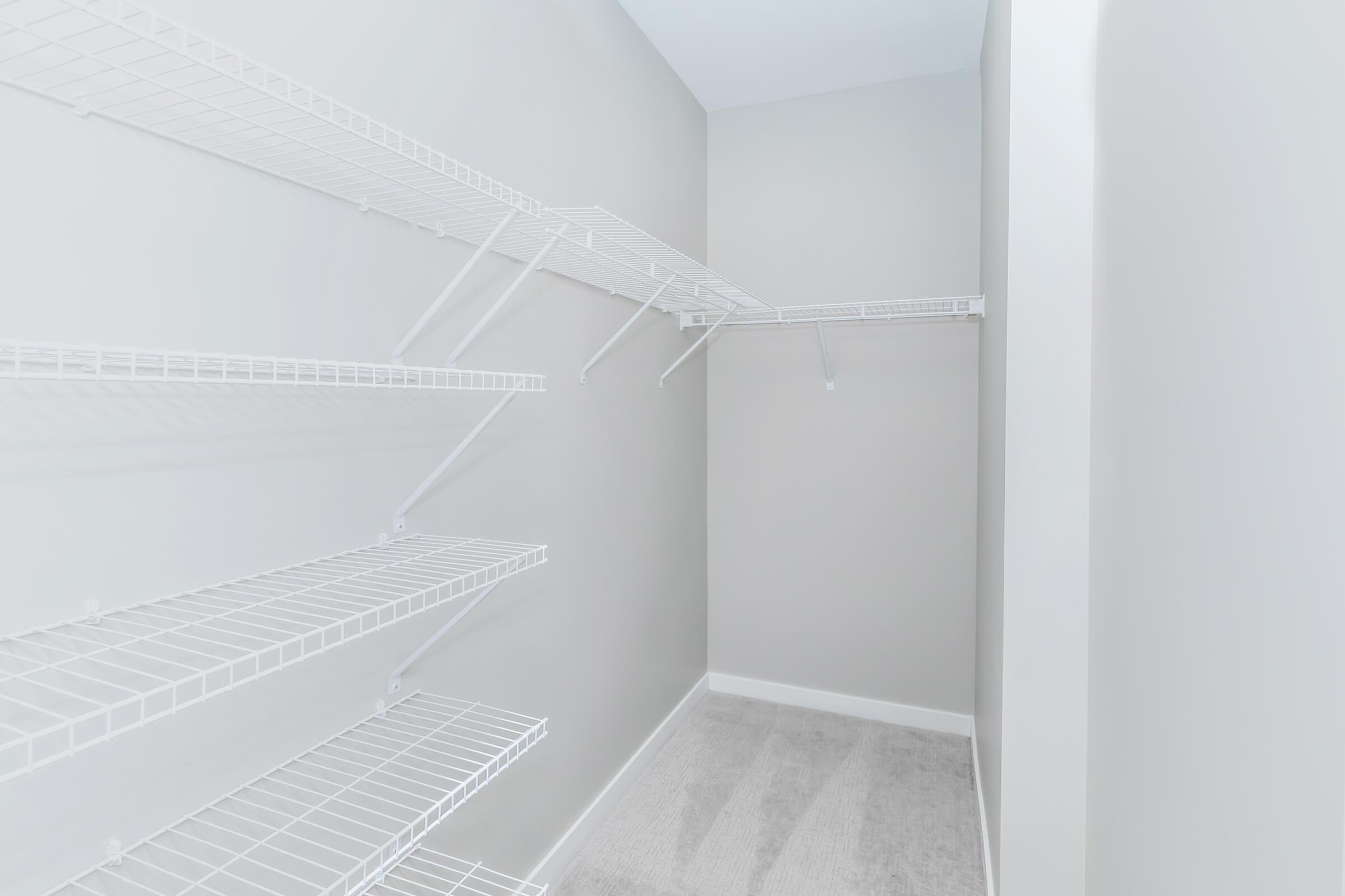 Empty closet with light gray walls and white wire shelving. The shelves are arranged on two walls, creating a spacious and organized storage area. The floor is carpeted in a light color, enhancing the bright and clean appearance of the room.