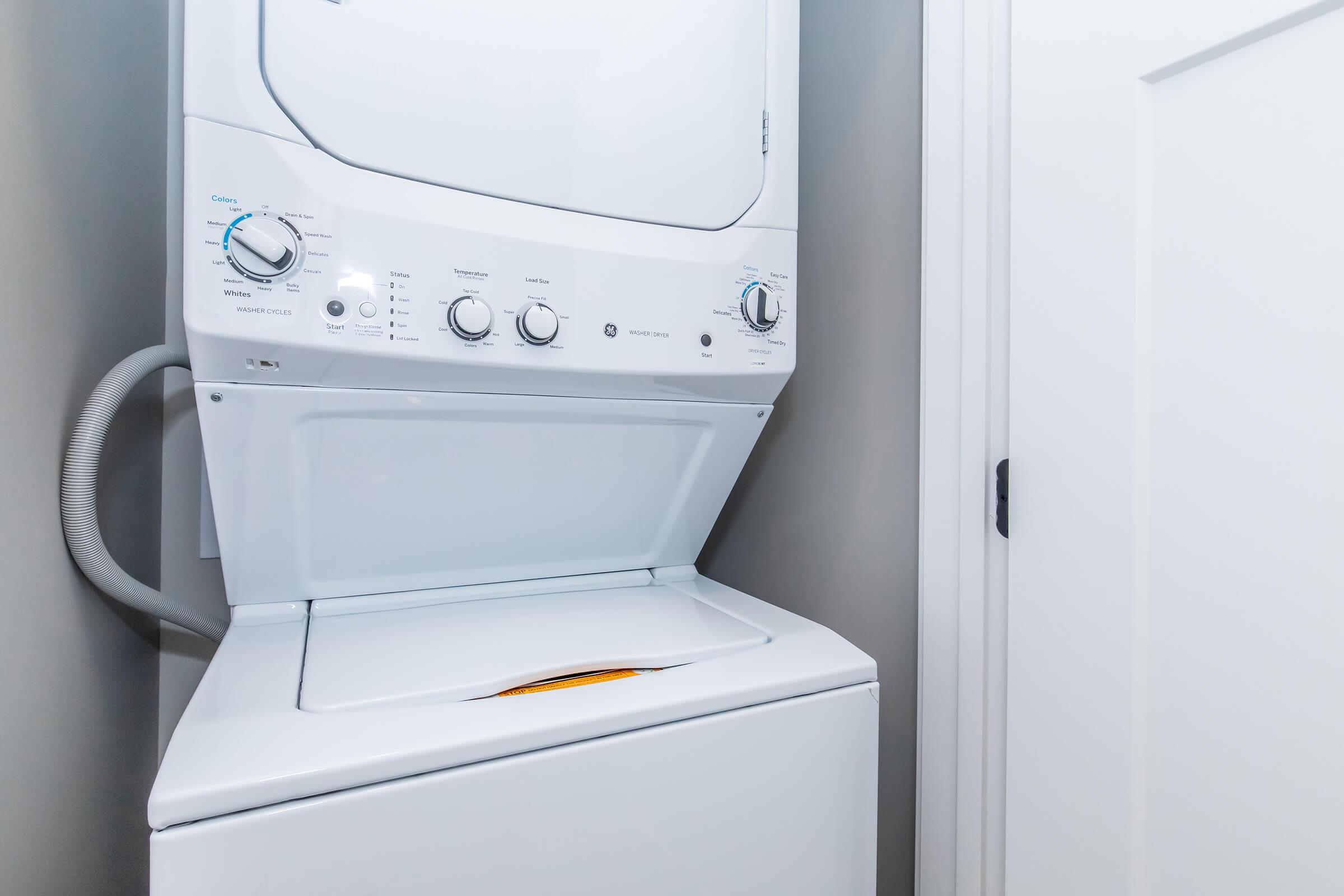 A stacked washer and dryer located in a small laundry area. The unit features a control panel with various knobs and settings, and the dryer is positioned above the washing machine. The surrounding wall is a light color, and there is a closed white door nearby.