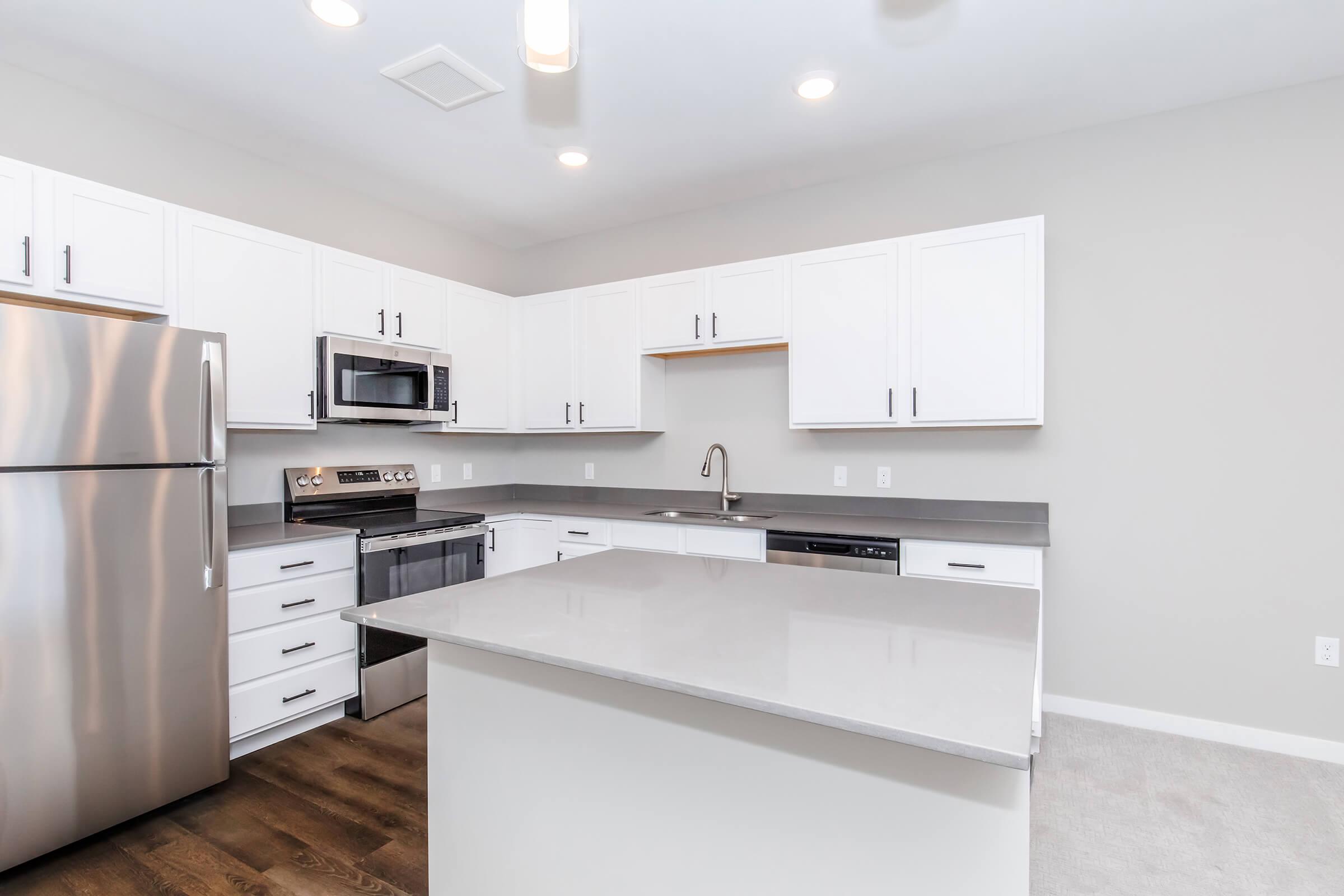 Modern kitchen featuring stainless steel appliances, including a refrigerator and microwave. White cabinets line the walls, complemented by a gray countertop and an island in the center. The space is illuminated by overhead lights, with a light gray wall color and stylish hardwood flooring.