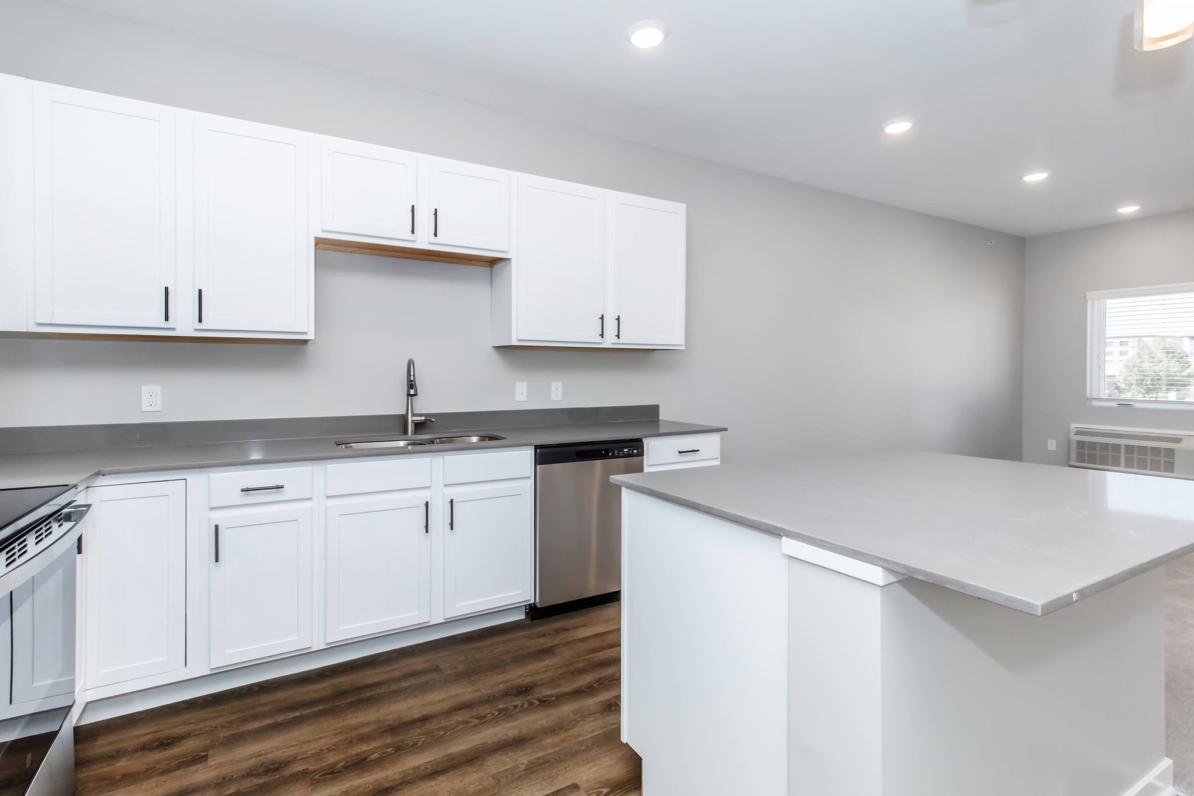 Modern kitchen featuring white cabinetry, stainless steel appliances, and a sleek countertop. The open layout includes a spacious island and neutral-colored walls, with natural light from a nearby window. The flooring is wooden, adding warmth to the contemporary design.