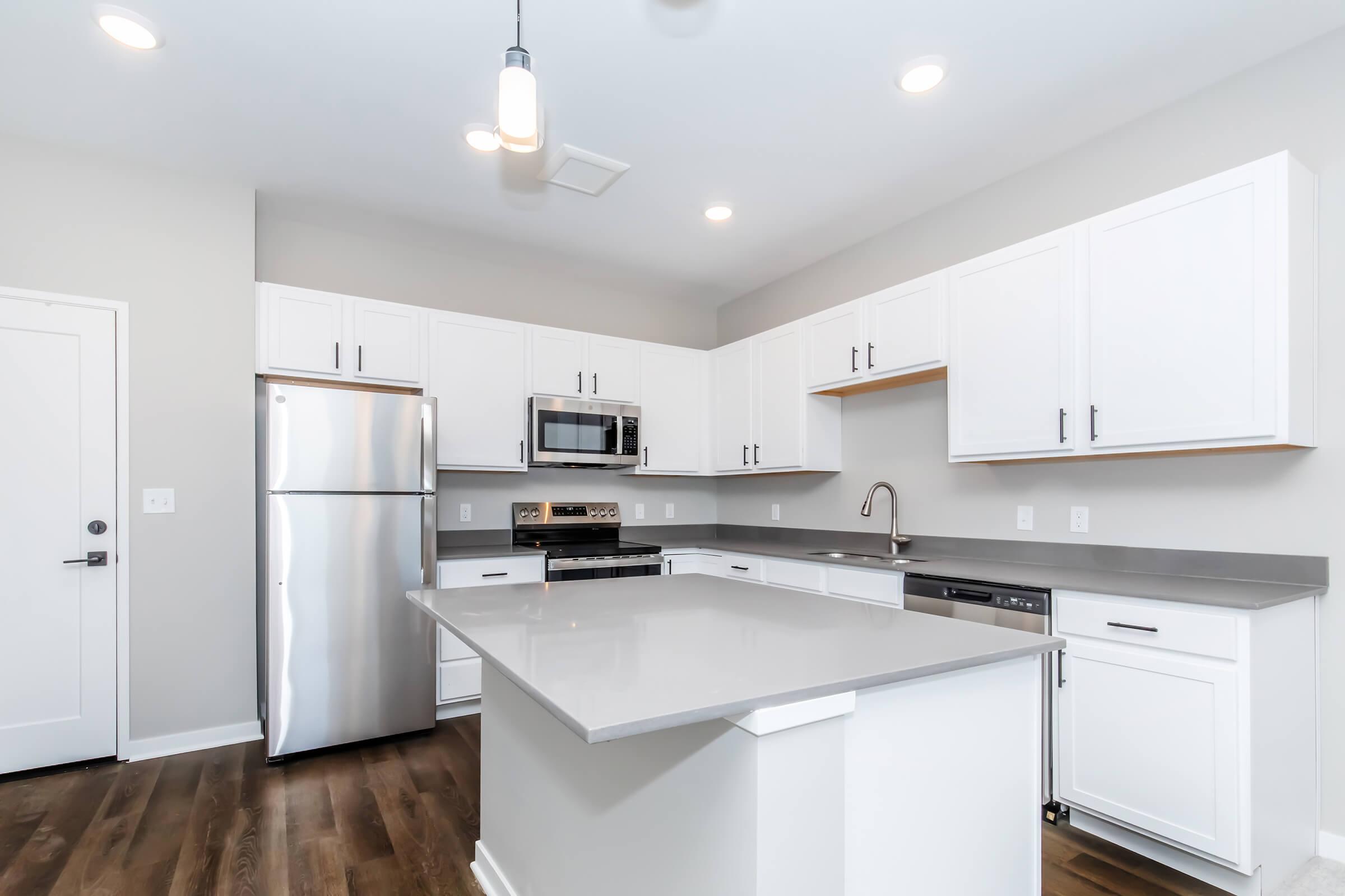 Modern kitchen with white cabinetry, stainless steel appliances including a refrigerator and microwave, a gray countertop, and a central island. Light fixtures illuminate the space, and there is a door leading to another area. The walls are painted in a soft gray, creating a clean and contemporary look.