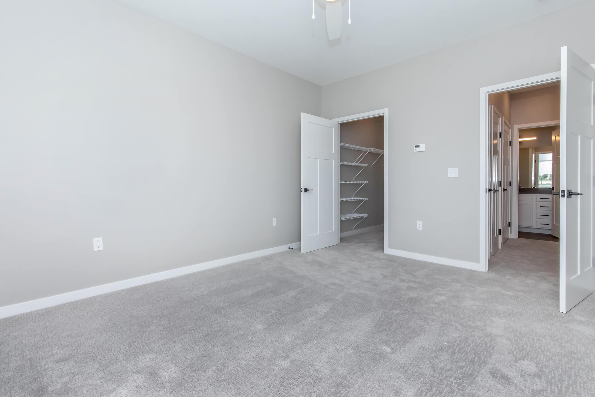 A spacious, empty room featuring light gray walls and plush carpet. There are two open doors: one leads to a closet with shelving, and the other to a bathroom area. The room is well-lit, showcasing a ceiling fan and a clean, modern design.