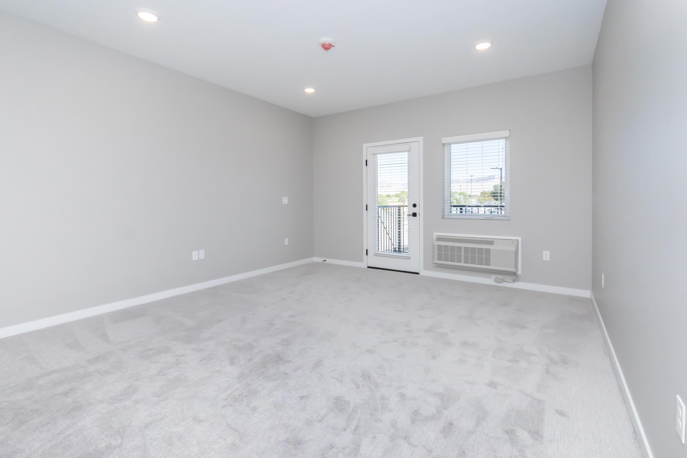 A spacious, empty room with light gray walls and plush carpet. It features a sliding glass door leading to a small balcony and a wall-mounted air conditioning unit. Soft natural light enters through the door's window, creating a bright atmosphere.