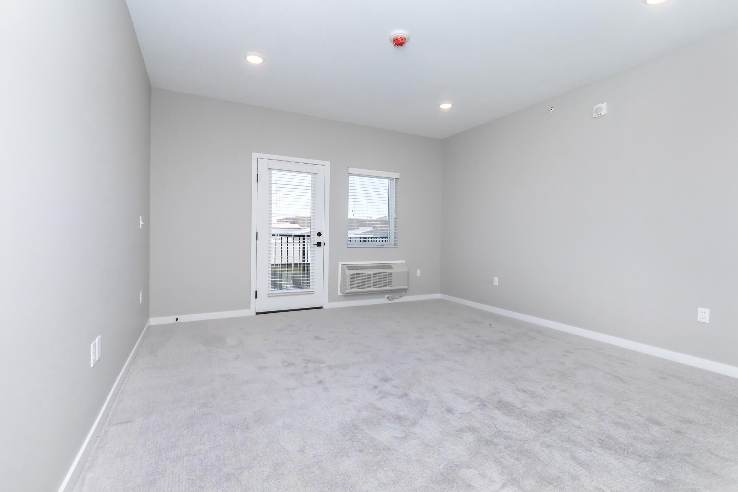A light gray room with freshly painted walls and carpet. It features two small windows, a sliding door leading to a balcony, and an air conditioning unit on the wall. The space is empty, providing a clean and spacious feel, suitable for furnishing.