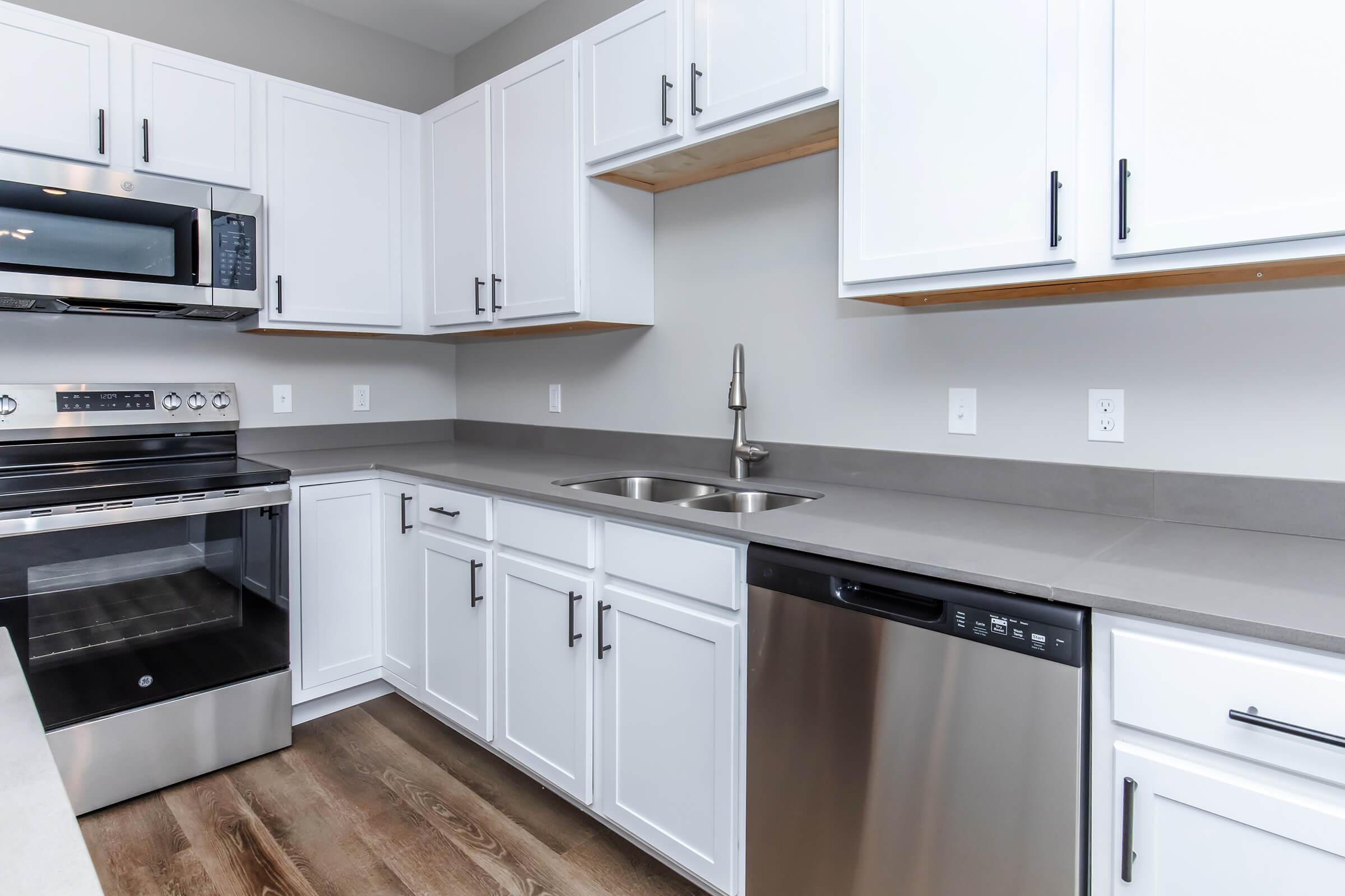 Modern kitchen featuring stylish white cabinets with black handles, a stainless steel microwave, electric stove, and dishwasher. The countertop is gray, complemented by a sleek sink and minimalist decor. Natural light brightens the space, creating a warm and inviting atmosphere.