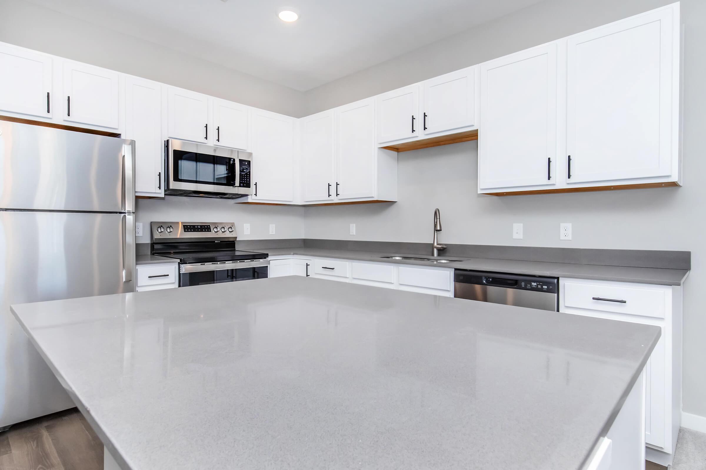 Modern kitchen featuring white cabinets, stainless steel appliances, and a large gray countertop. The space includes a refrigerator, microwave, oven, and dishwasher, with a clean and minimalist design. Natural light illuminates the area, highlighting the sleek finishes and organized layout.