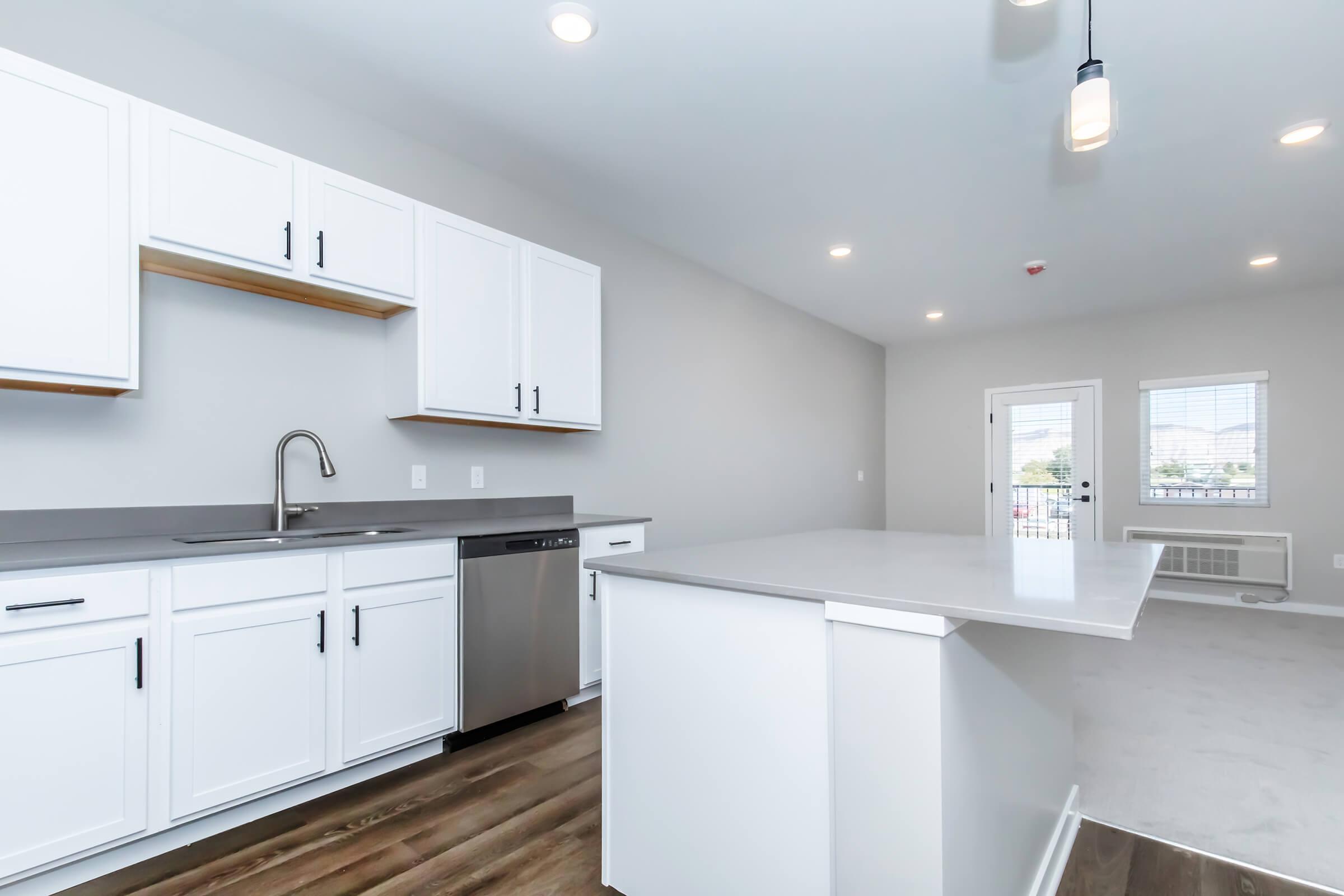 Contemporary kitchen with white cabinets, stainless steel dishwasher, and a gray countertop. Open layout features a kitchen island, modern light fixtures, and light-colored walls. A window and door lead to an outdoor area, creating a bright and airy atmosphere.