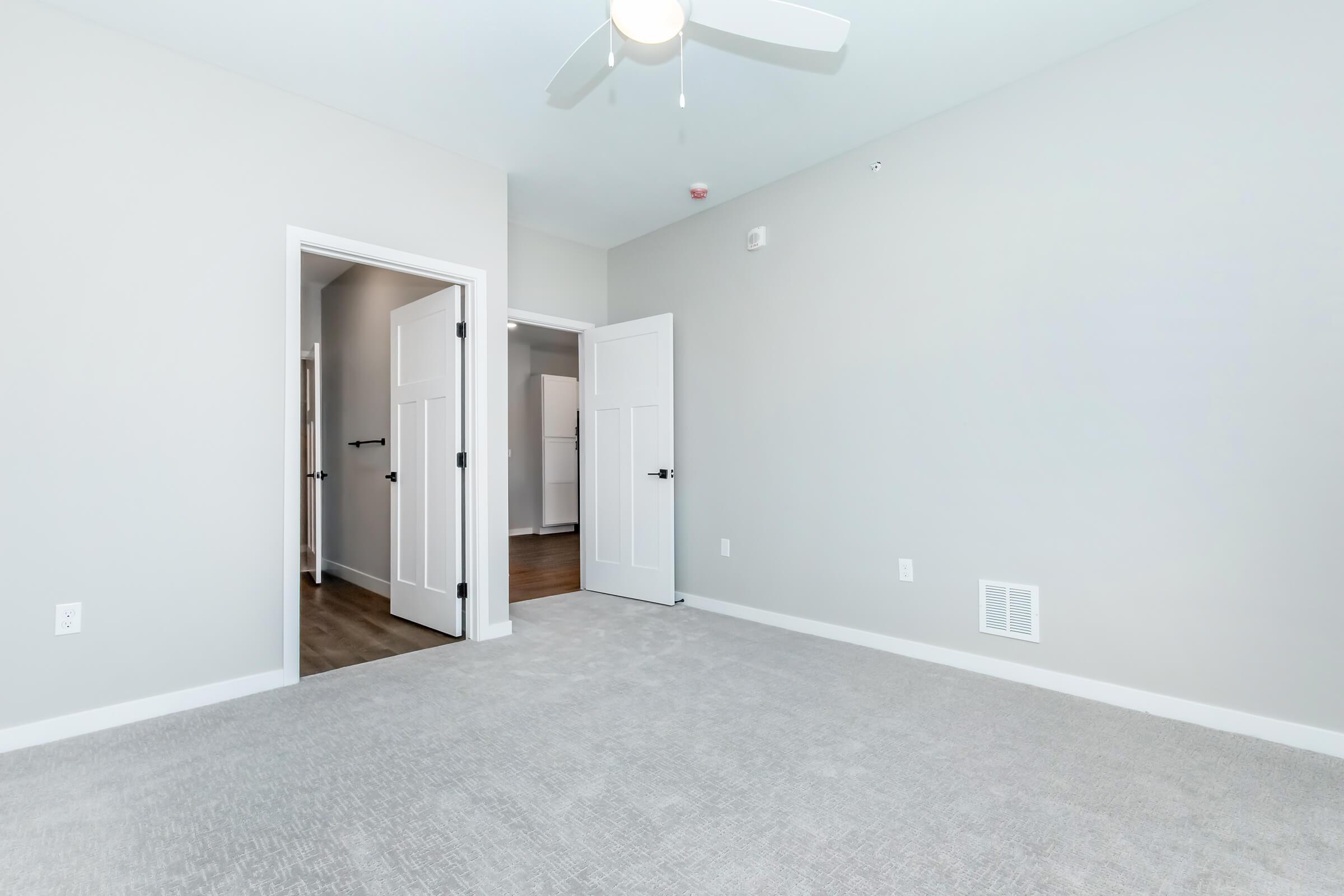 A spacious, empty room featuring light gray walls and plush carpeting. Two open doorways lead to adjacent rooms, while a ceiling fan hangs above. The room has a modern design with minimal decor, providing a clean and bright atmosphere.