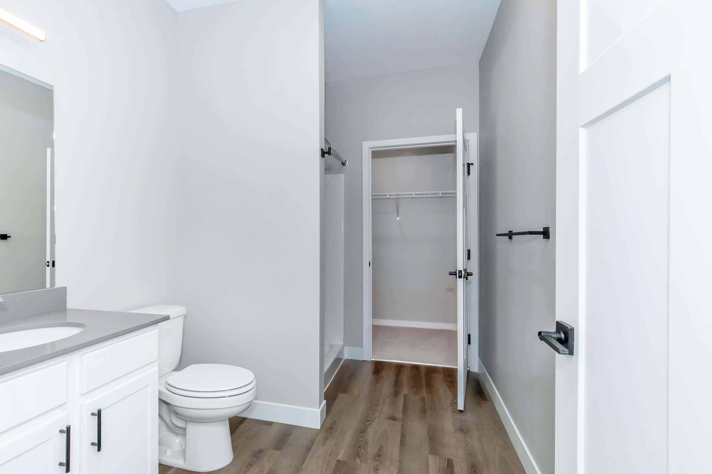 Modern bathroom featuring a white toilet, a double sink vanity with gray countertops, and light wood flooring. A door leads to a closet area with a hanging rod, while another door opens to the hallway. The walls are painted in a soft gray color, providing a clean and contemporary look.