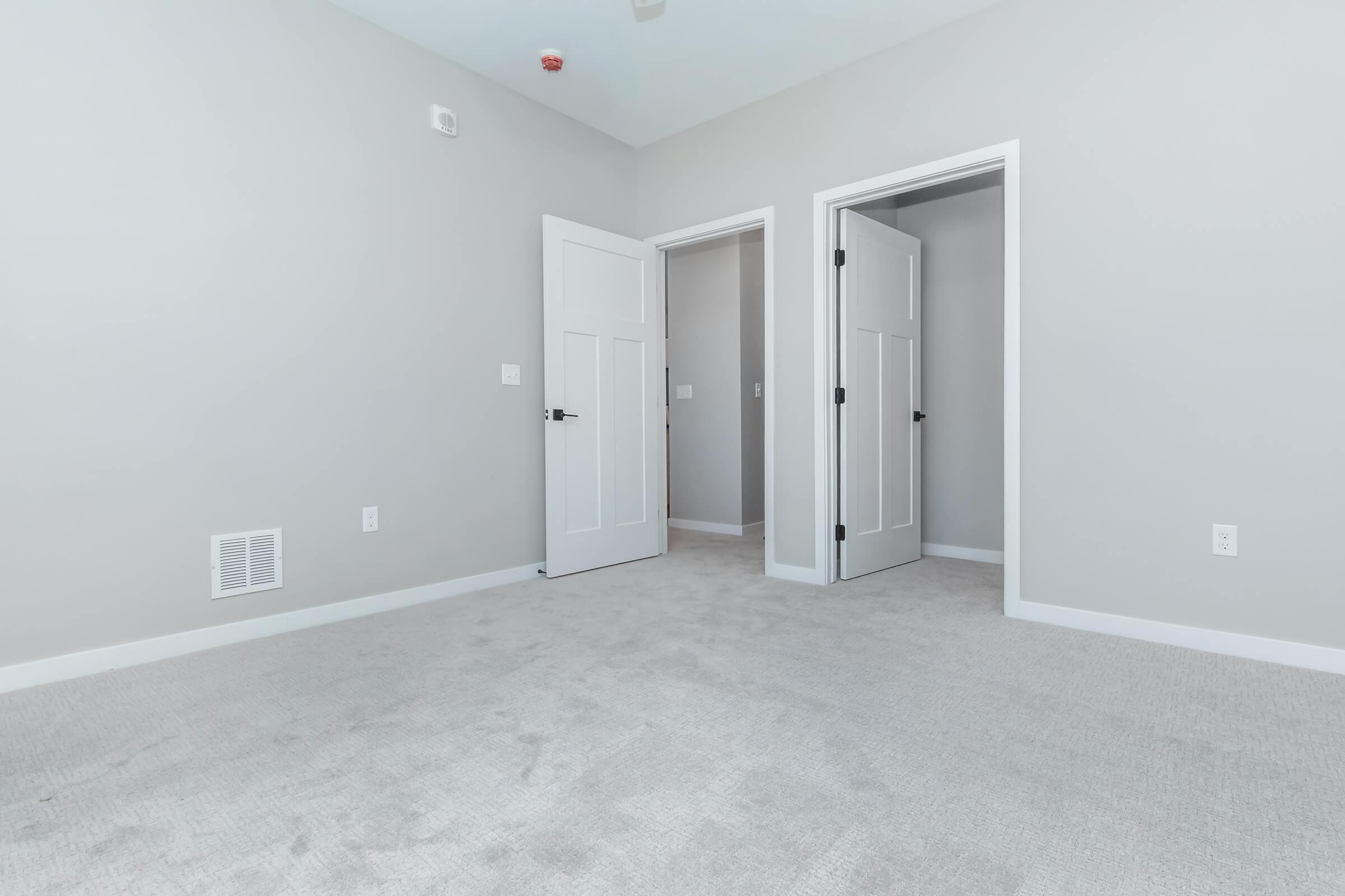Interior view of a light gray room with carpeted floor, featuring two open white doors leading to adjacent rooms. The walls are painted in a similar light gray shade, and there is a vent on the wall. The space appears spacious and well-lit, suitable for various arrangements.