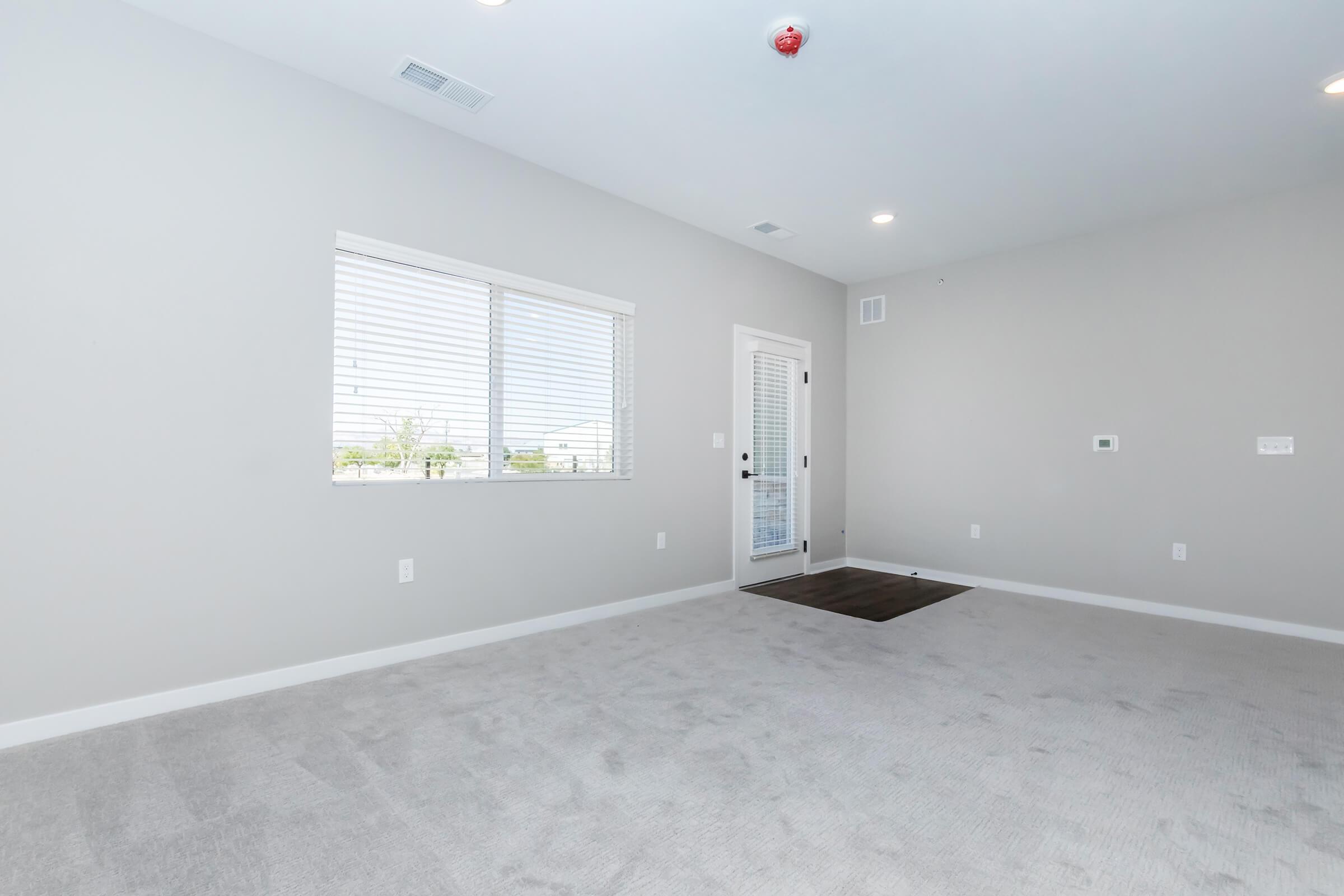 A bright, empty room with light gray walls and carpet. There is a window with white blinds on one wall, allowing natural light to enter. A door leads outside, and there are recessed lights on the ceiling. The space is clean and minimal, ready for furnishing.