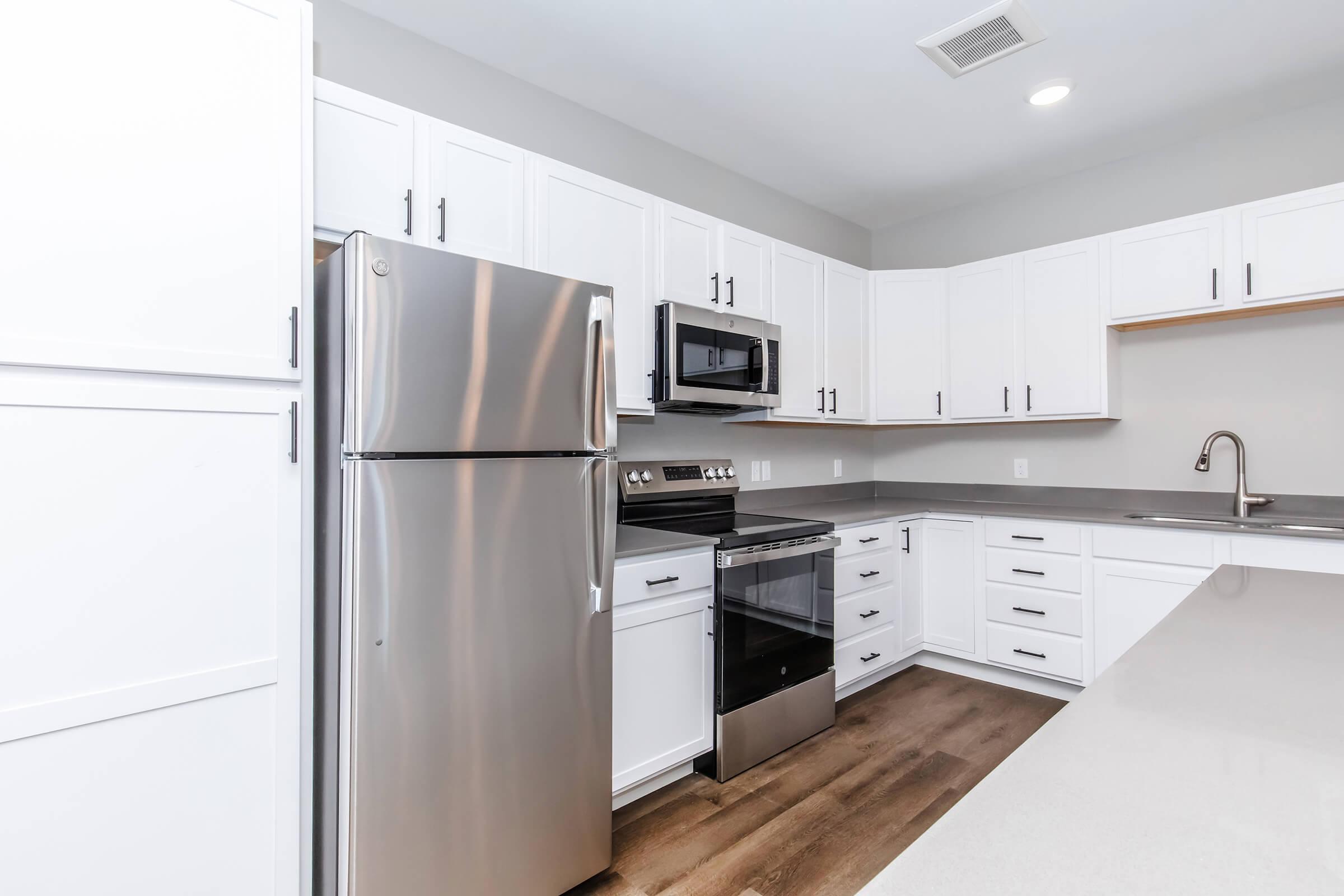 Modern kitchen featuring stainless steel appliances including a refrigerator, oven, and microwave. The cabinetry is white with silver hardware, and the countertops are light-colored. The floor has a wooden finish, creating a clean and contemporary look.