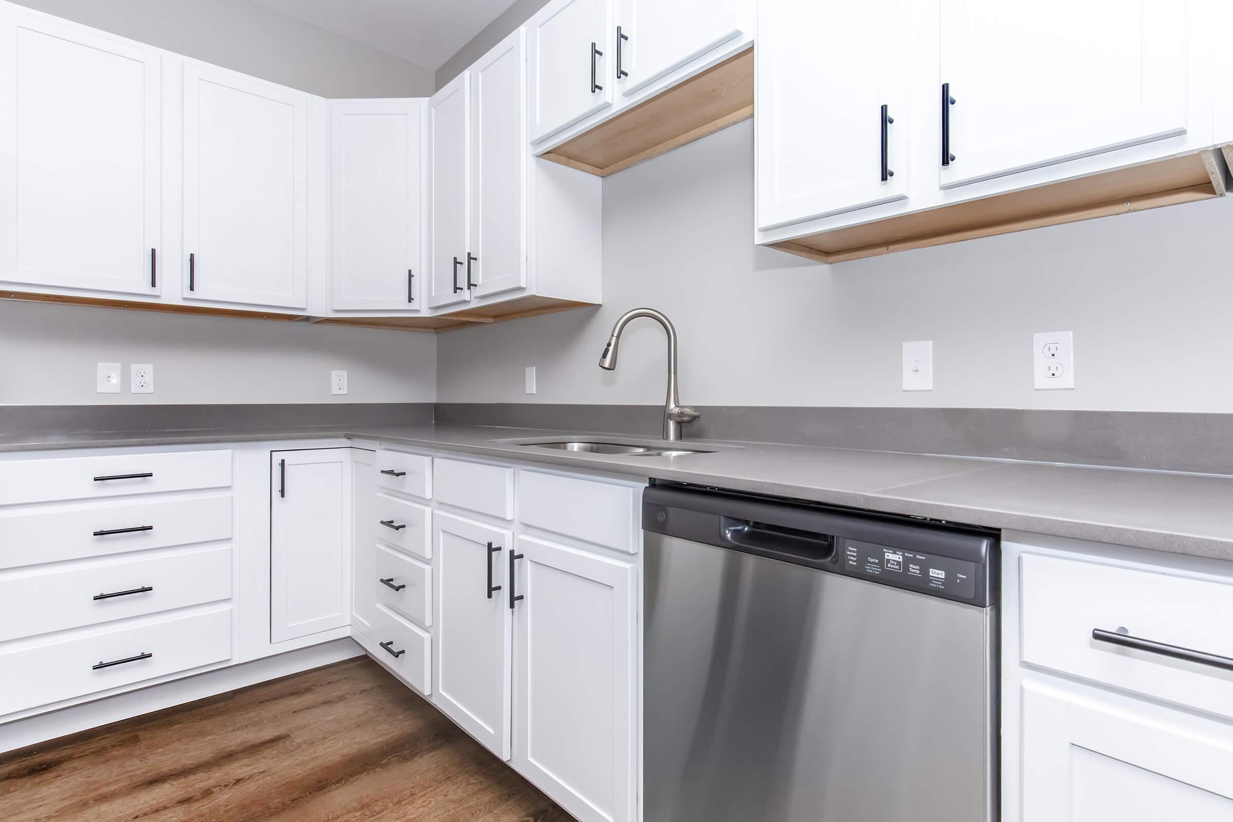 Modern kitchen with white cabinets and drawers, gray countertops, and stainless steel appliances, including a dishwasher. The design features a minimalist aesthetic with a contemporary faucet and open shelving above. Hardwood flooring completes the stylish look.
