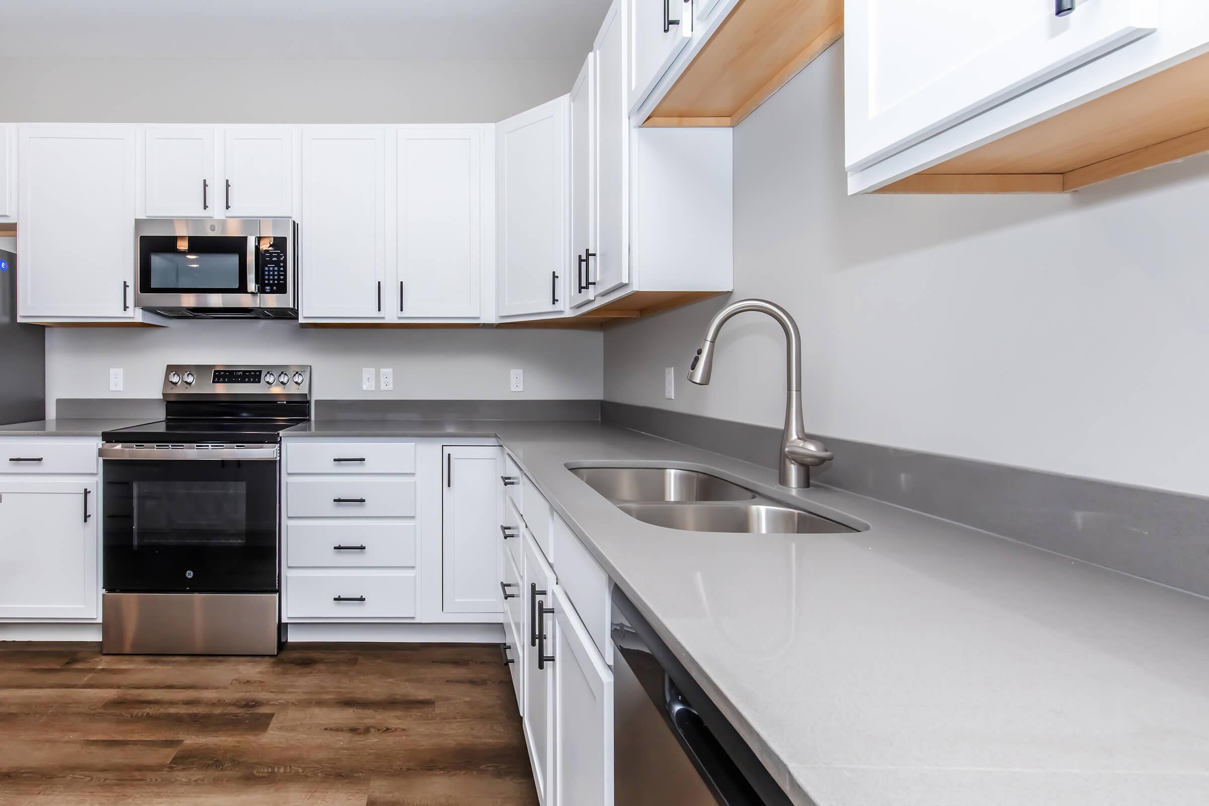 Modern kitchen featuring white cabinetry, stainless steel appliances including a microwave and oven, a double sink, and sleek gray countertops. The design highlights clean lines and a spacious layout with wooden flooring.