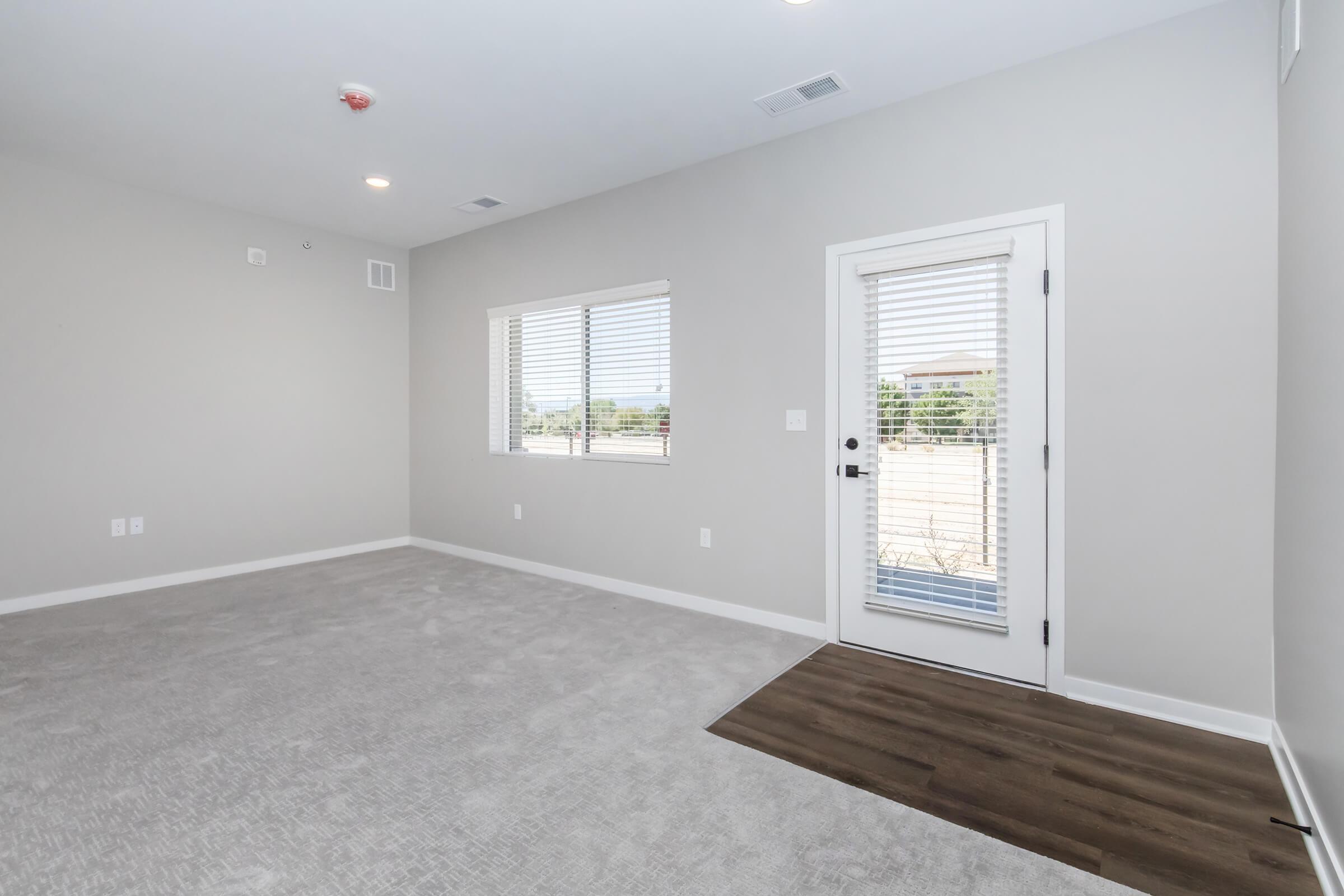 A spacious room with light gray walls and carpet, featuring a large window with horizontal blinds and a glass door leading outside. The flooring near the door is hardwood, contrasting with the carpet. The room appears bright and empty, suitable for various uses.